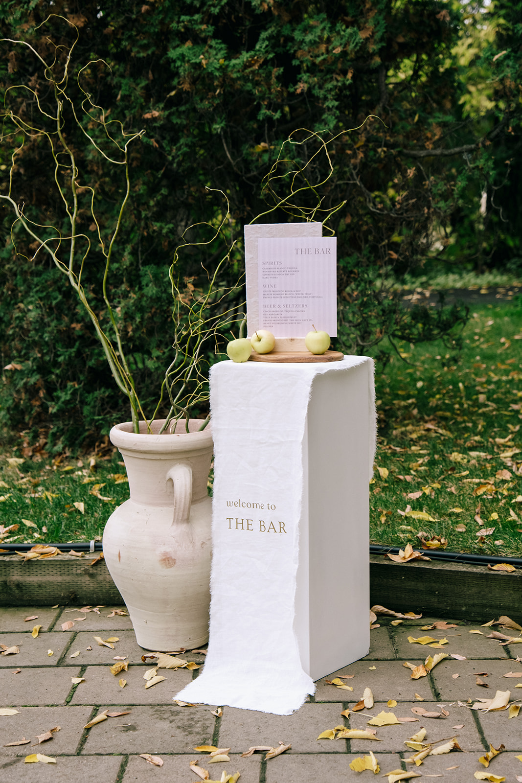 Pedestal bar stand sign design layered with flowy fabric, organic neutral vases with grasses and green apples for a cottage core wedding.