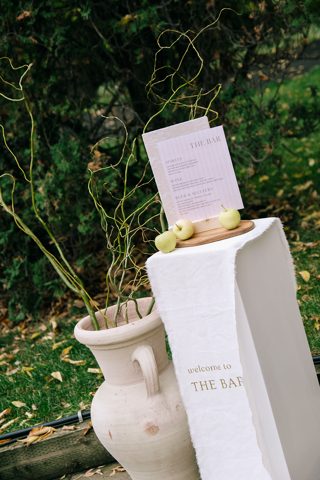 Pedestal bar stand sign design layered with flowy fabric, organic neutral vases with grasses and green apples for a cottage core wedding.