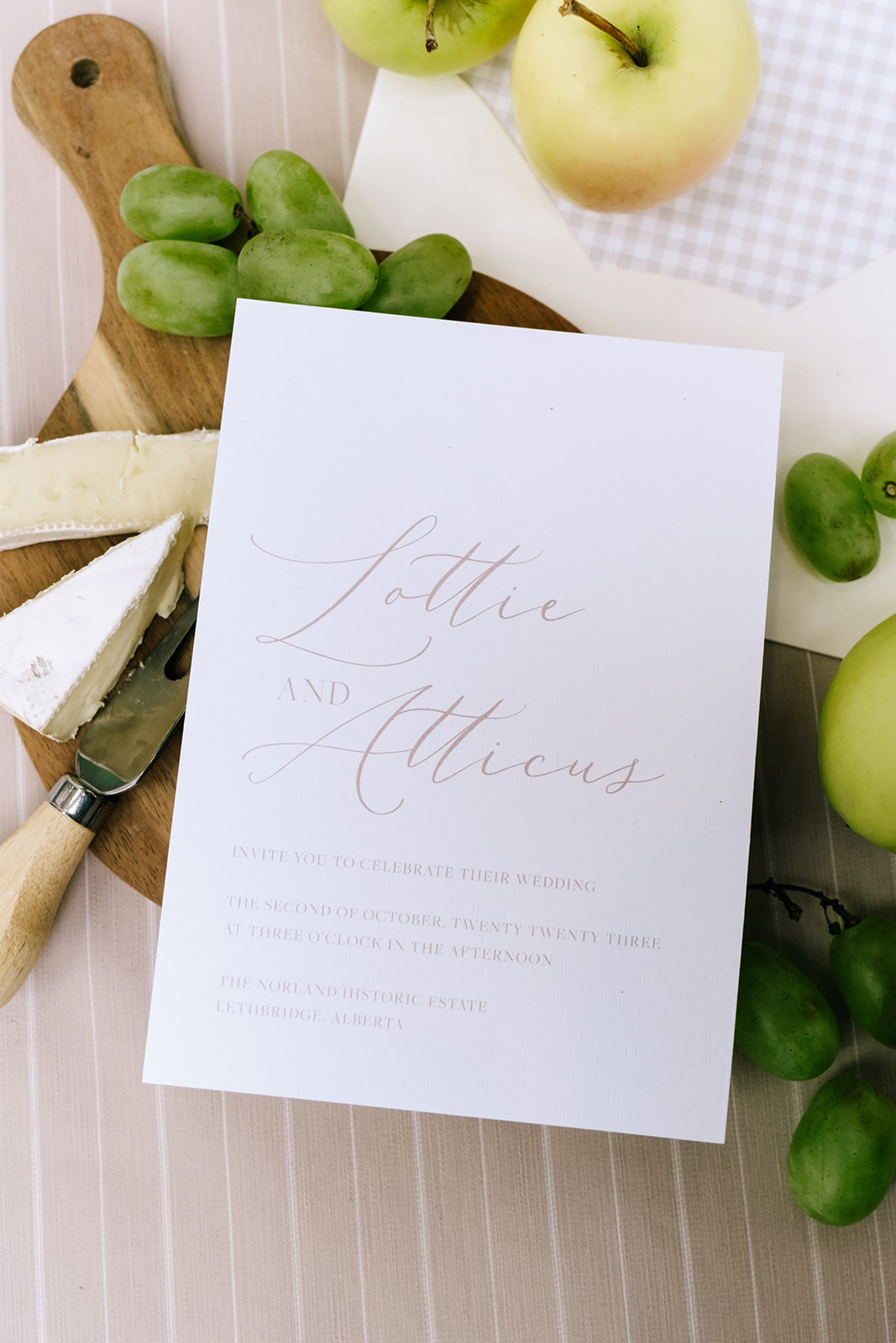 Close-up of wedding invitation flatlay surrounded by fruit and cheese on a wood cutting board, embracing the cottage core aesthetic.