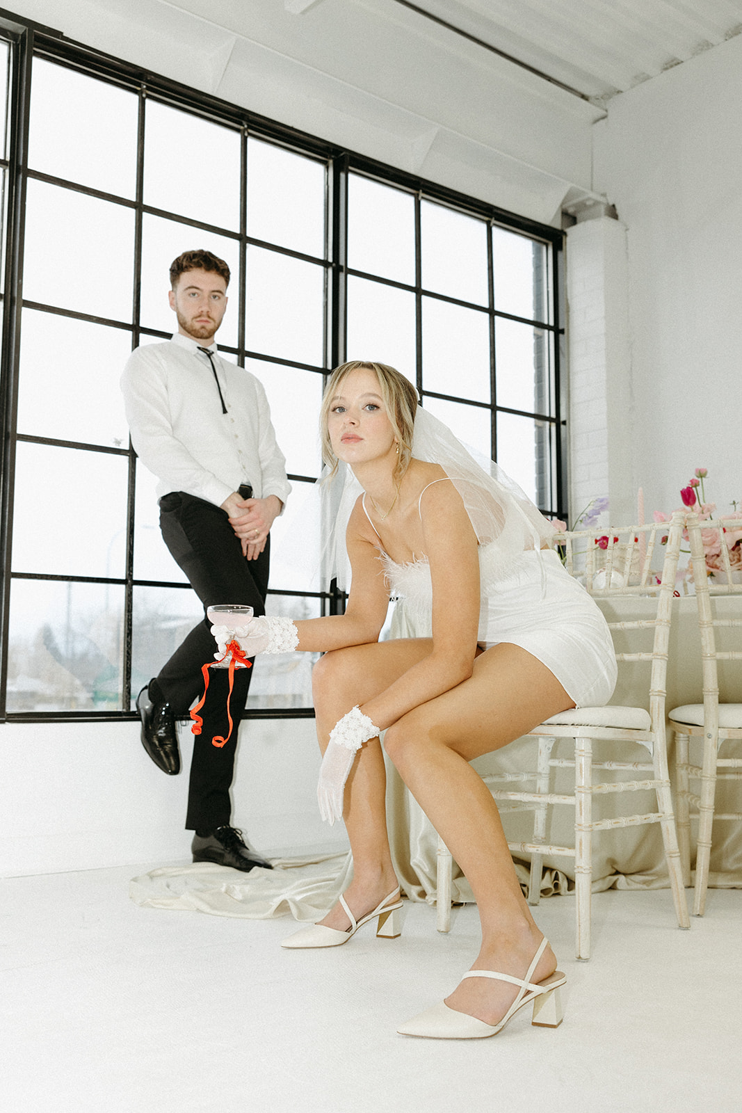 Bride and groom in retro-inspired wedding attire, posed for a funky, creative wedding editorial photoshoot. Bride wearing short dress with feather details and vintage bridal gloves, holding champagne glass tied with red ribbon. 
