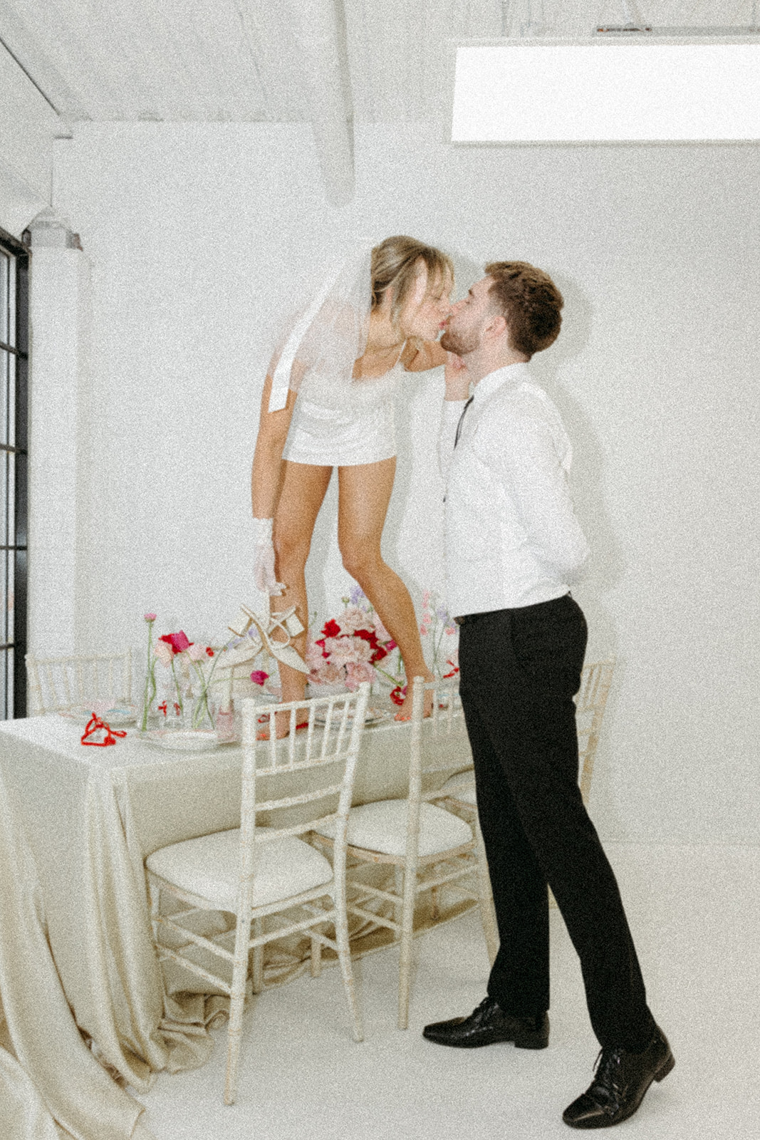 Editorial wedding shoot featuring retro wedding dress and groom’s suit, styled with modern minimalist elements. Funky reception setup with vibrant retro-inspired decor and playful design elements, bold coloured candles, and modern flowers. Bride and groom embracing a fun, unconventional pose in a funky retro setting.