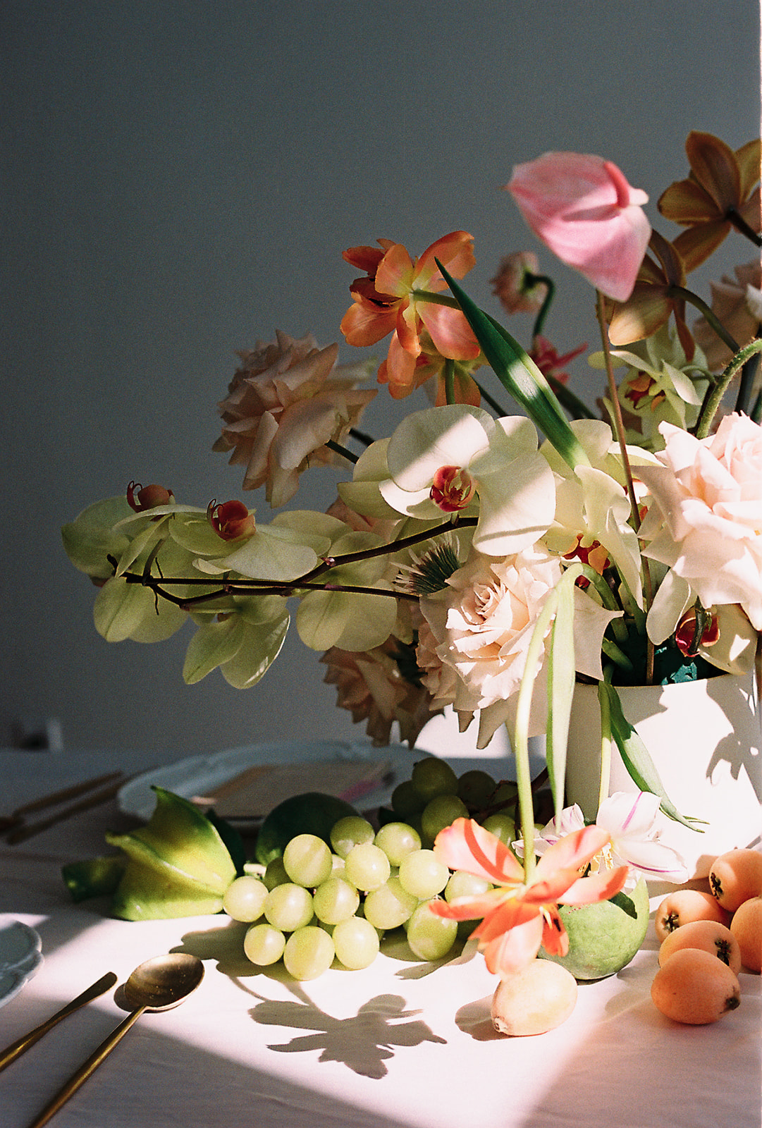 A vibrant close-up of a table arrangement featuring a lush display of flowers in soft peach, orange and cream tones. The floral arrangement is accompanied by fresh green grapes and apricots, adding a natural, elegant feel to the table setting. The lighting casts a soft warm glow over the arrangement, enhancing the colours and textures of the display. Fresh Fruit and Florals Wedding Table Centerpiece Ideas.