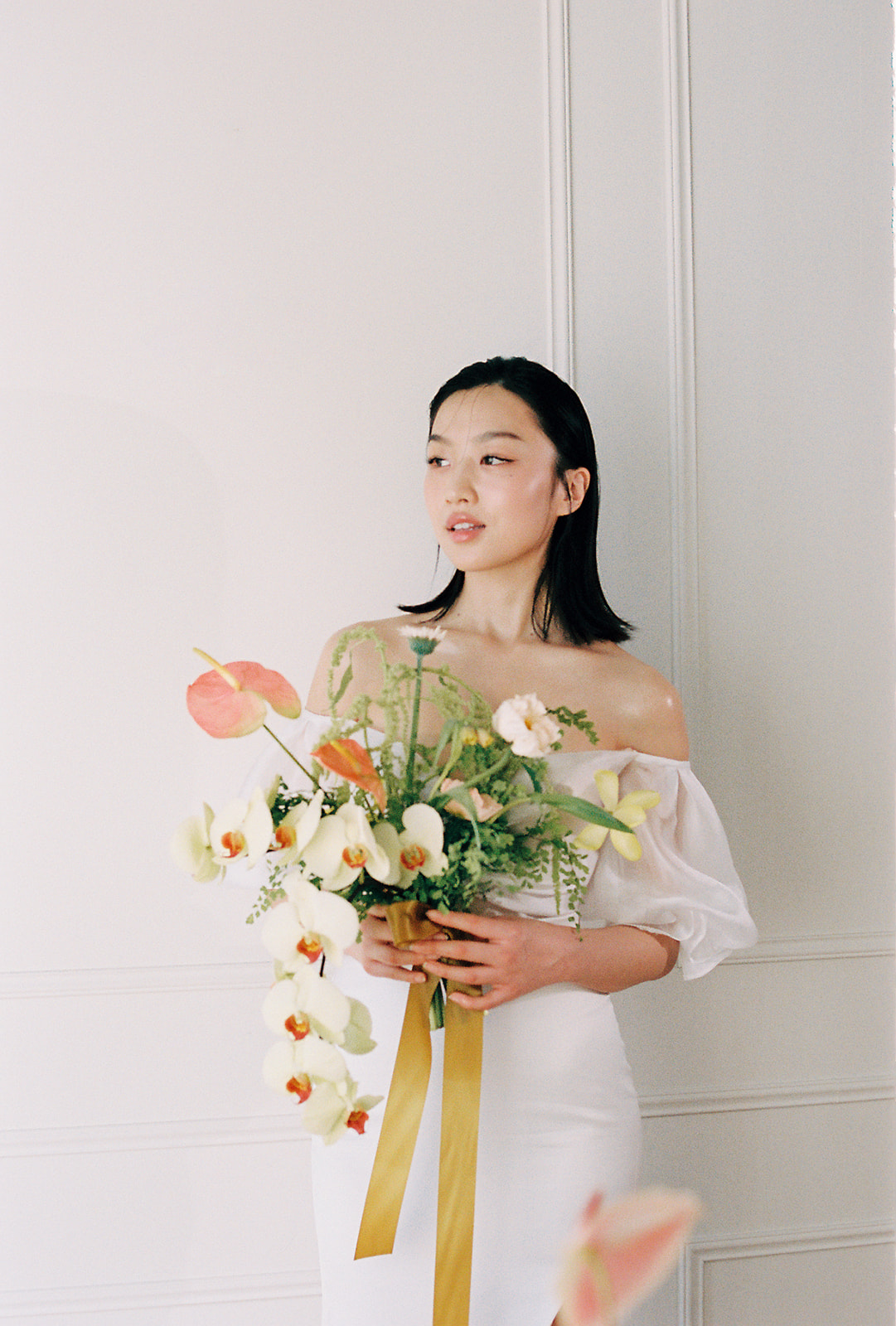 A bride in an elegant, off-the-shoulder white dress. She holds a bouquet of spring-inspired flowers, including peach-toned blooms and greenery. 