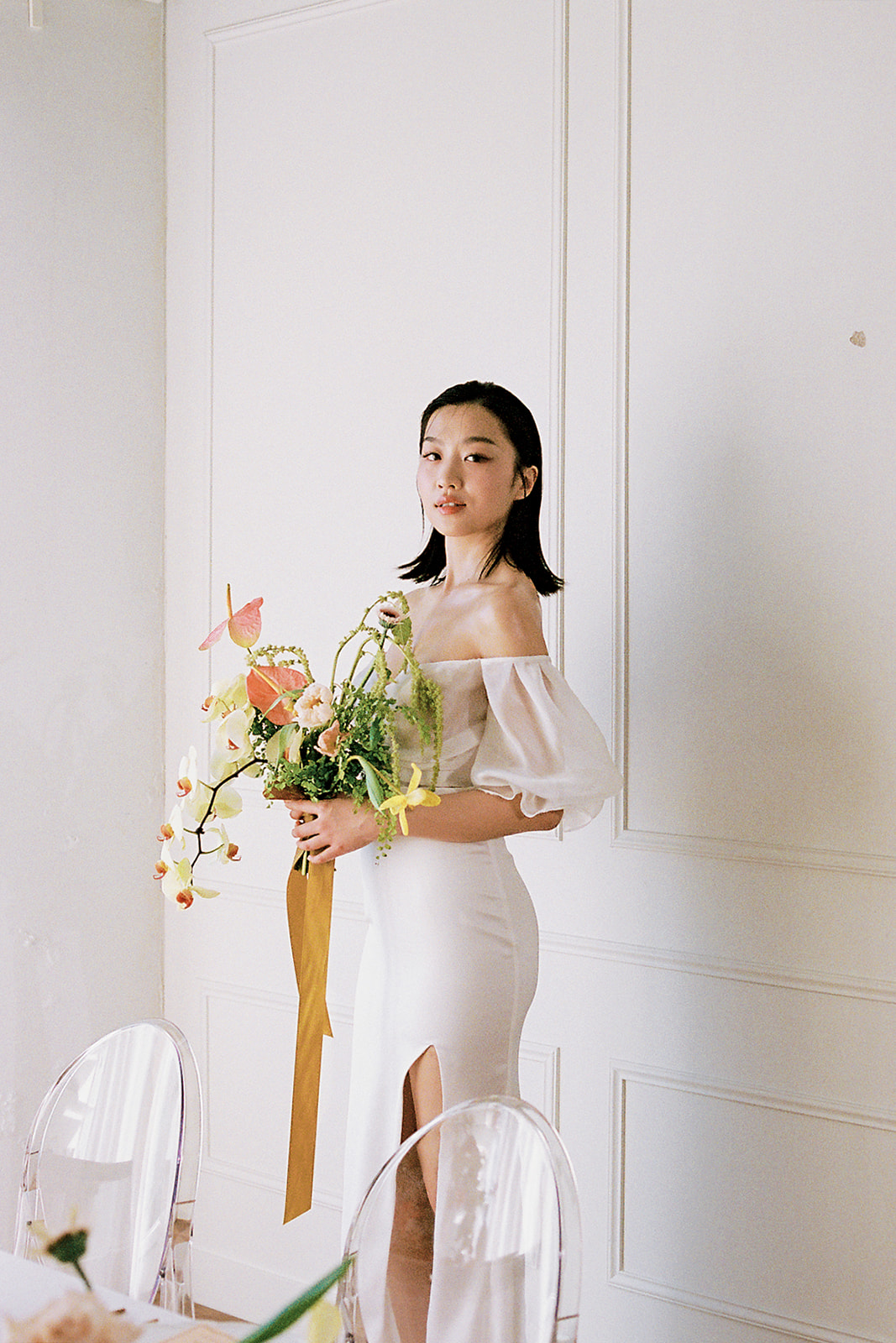 A bride in an elegant, off-the-shoulder white dress. She holds a bouquet of spring-inspired flowers, including peach-toned blooms and greenery. The room features minimalist decor with a beautifully set table in the foreground.