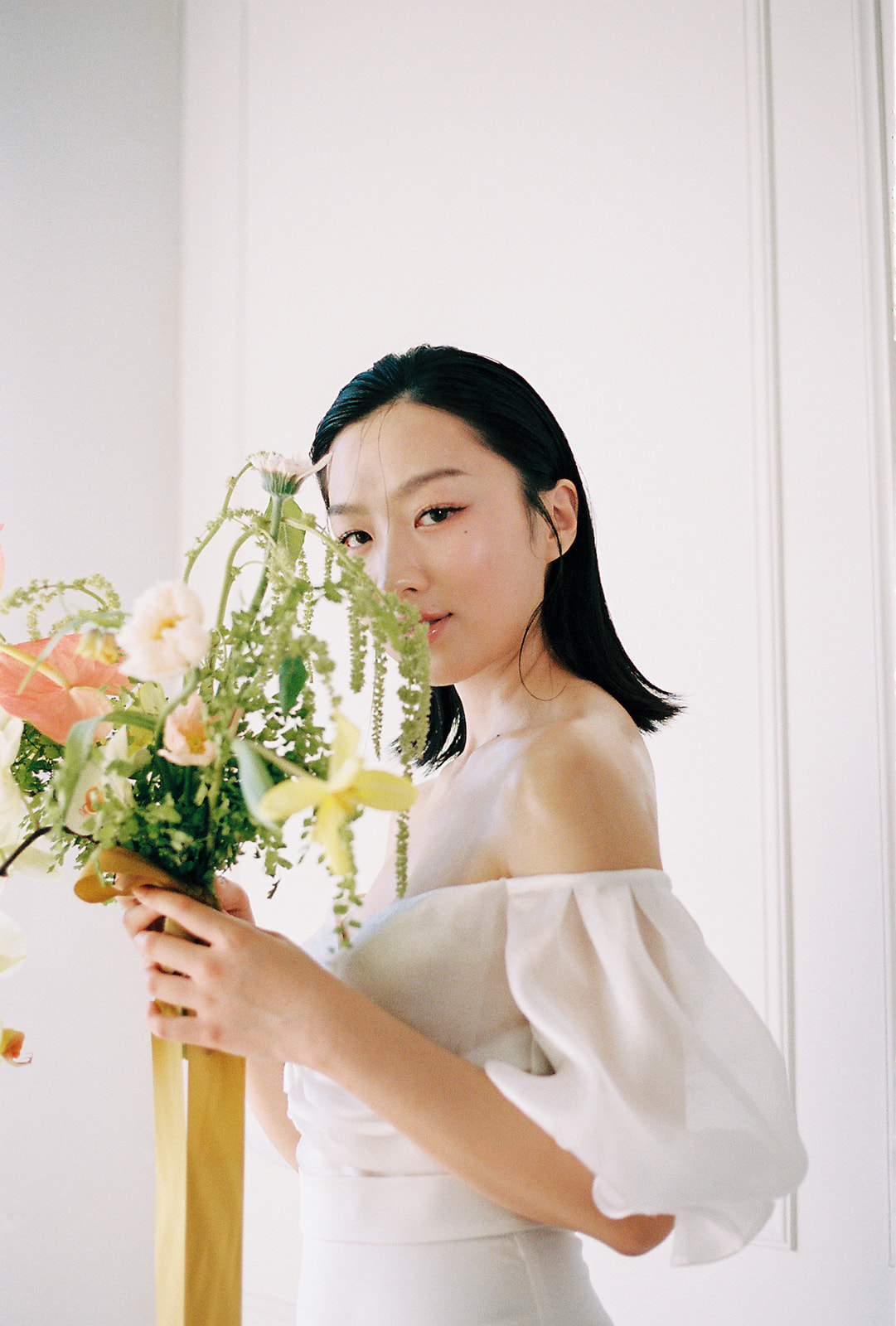 A bride in an elegant, off-the-shoulder white dress. She holds a bouquet of spring-inspired flowers, including peach-toned blooms and greenery, tied with a yellow silk ribbon.