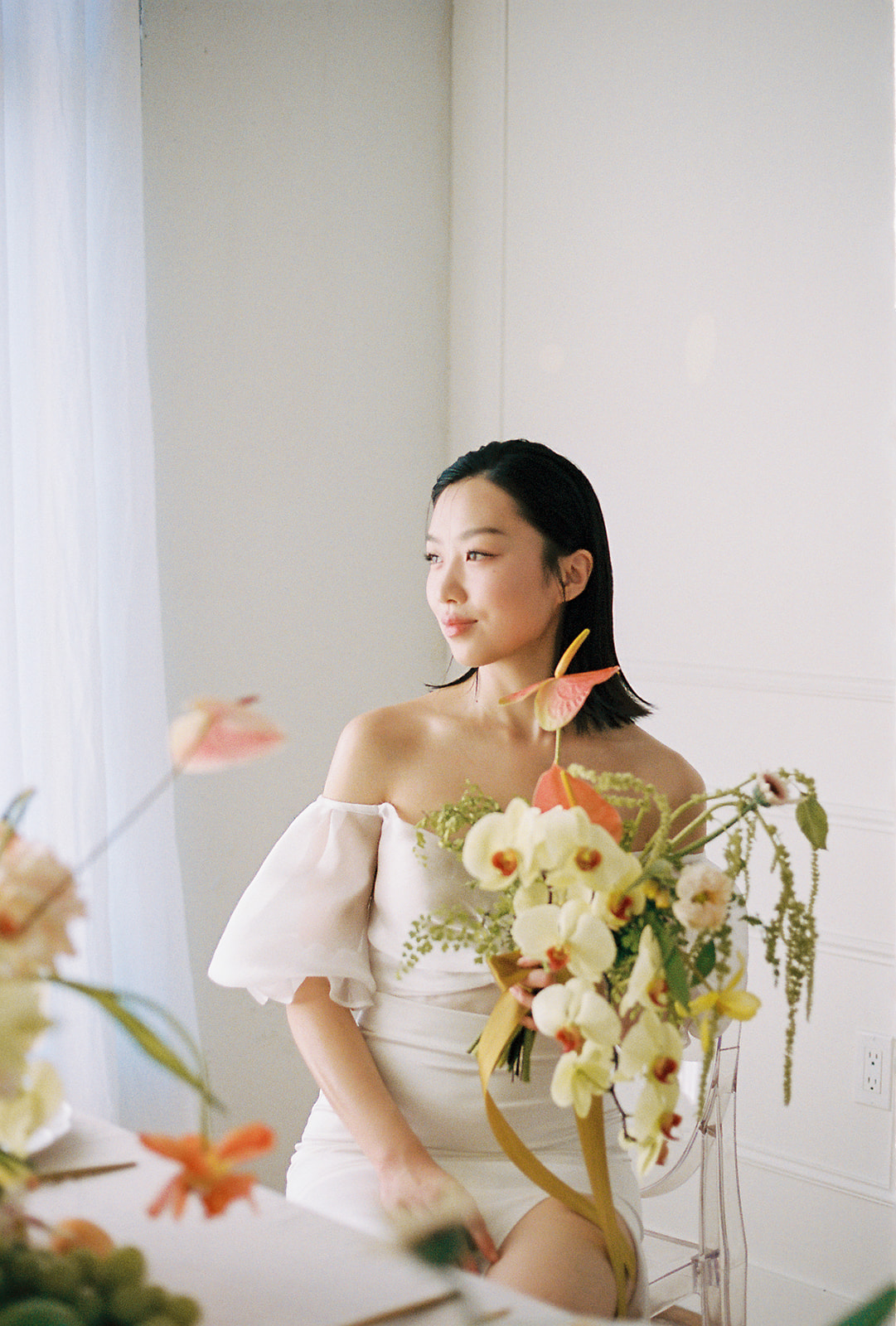 A bride in an elegant, off-the-shoulder white dress. She holds a bouquet of spring-inspired flowers, including peach-toned blooms and greenery, tied with a yellow silk ribbon.