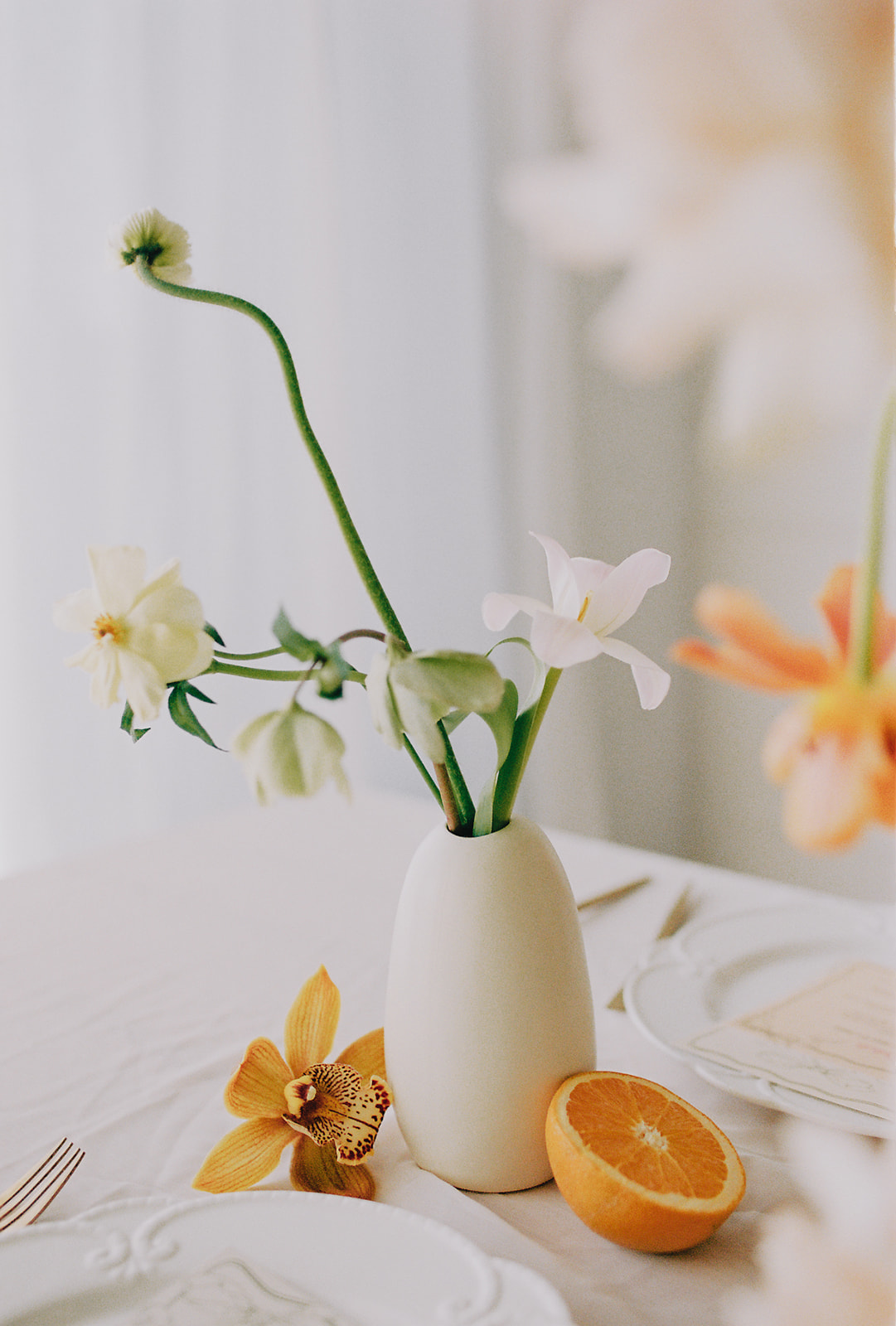 A vibrant close-up of a table arrangement featuring a ceramic bud vase and orange cut in half on a wedding tablescape. Peach, orange and cream wedding colour palette. Fruit and Florals Wedding