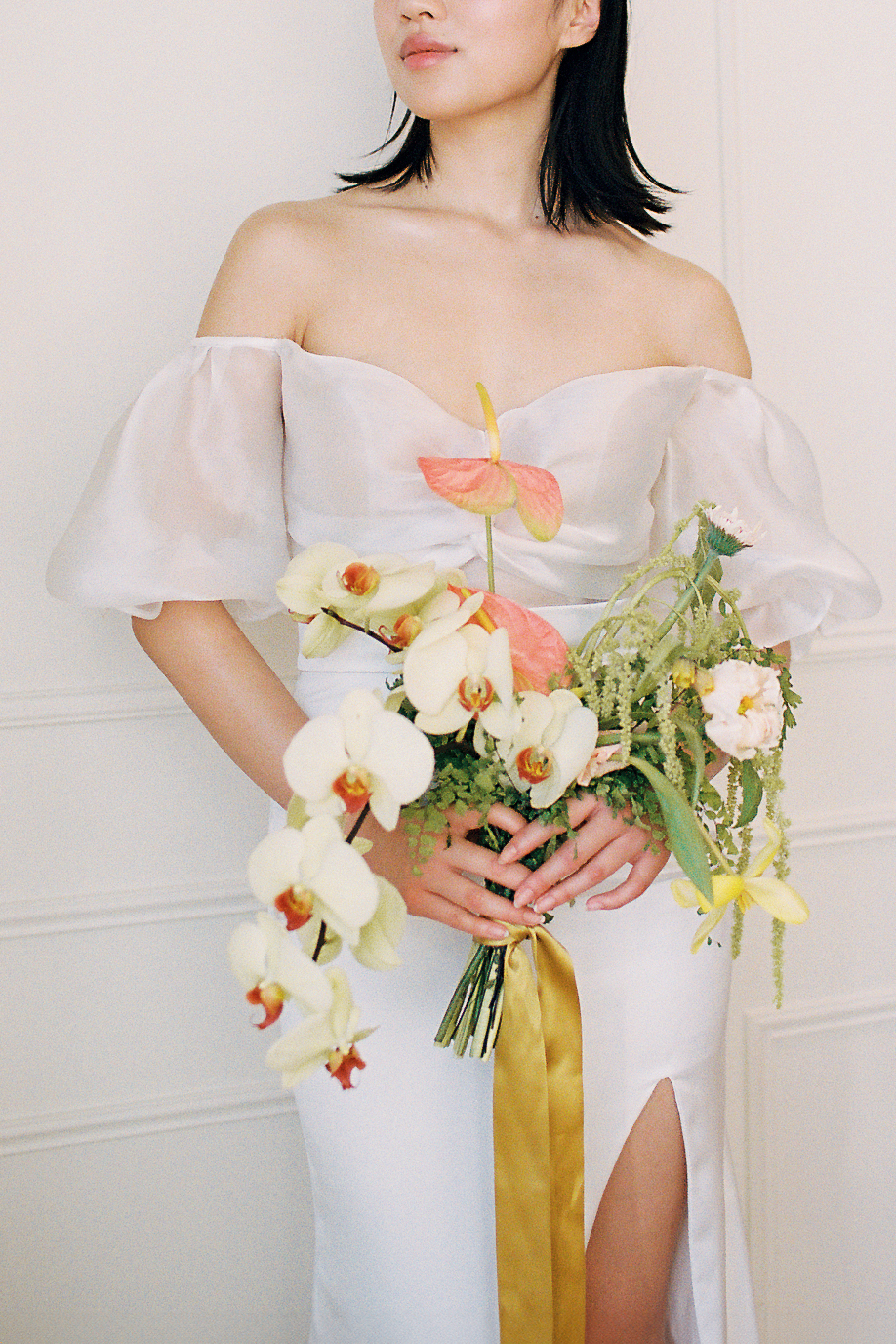 A bride in an elegant, off-the-shoulder white dress. She holds a bouquet of spring-inspired flowers, including peach-toned blooms and greenery, tied with a yellow silk ribbon. Citrus coloured wedding bouquet featured in this fresh fruit and florals wedding inspiration.