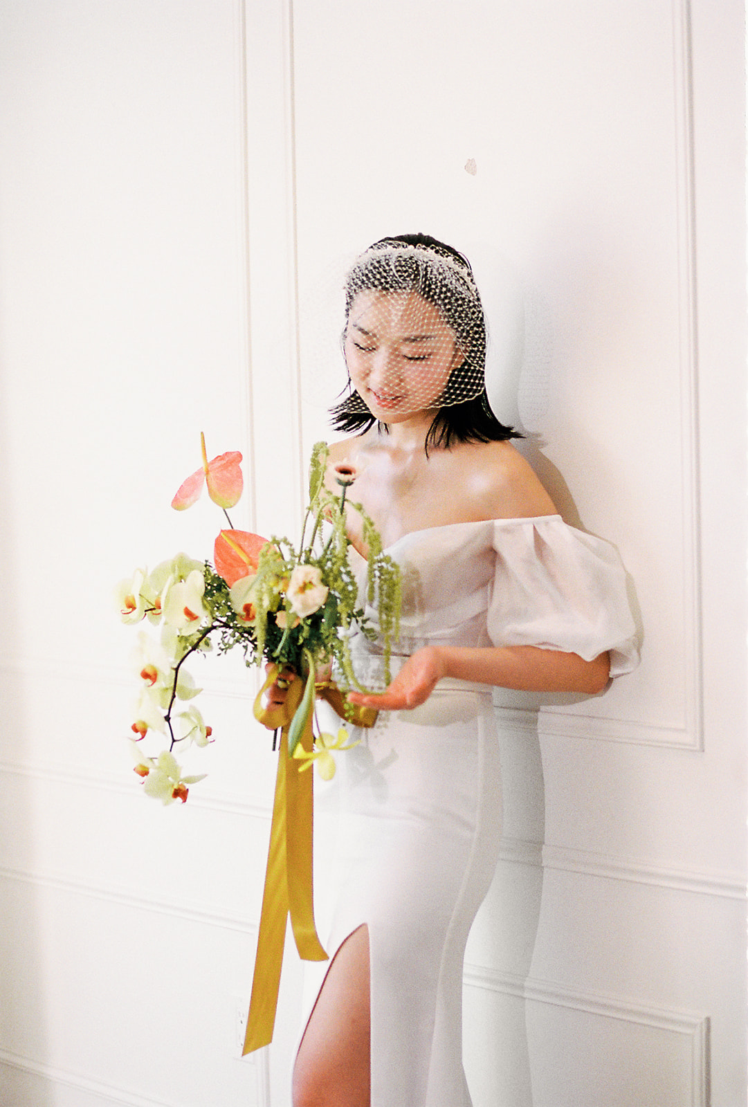 A bride in an elegant, off-the-shoulder white dress. She holds a bouquet of spring-inspired flowers, including peach-toned blooms and greenery, tied with a yellow silk ribbon.