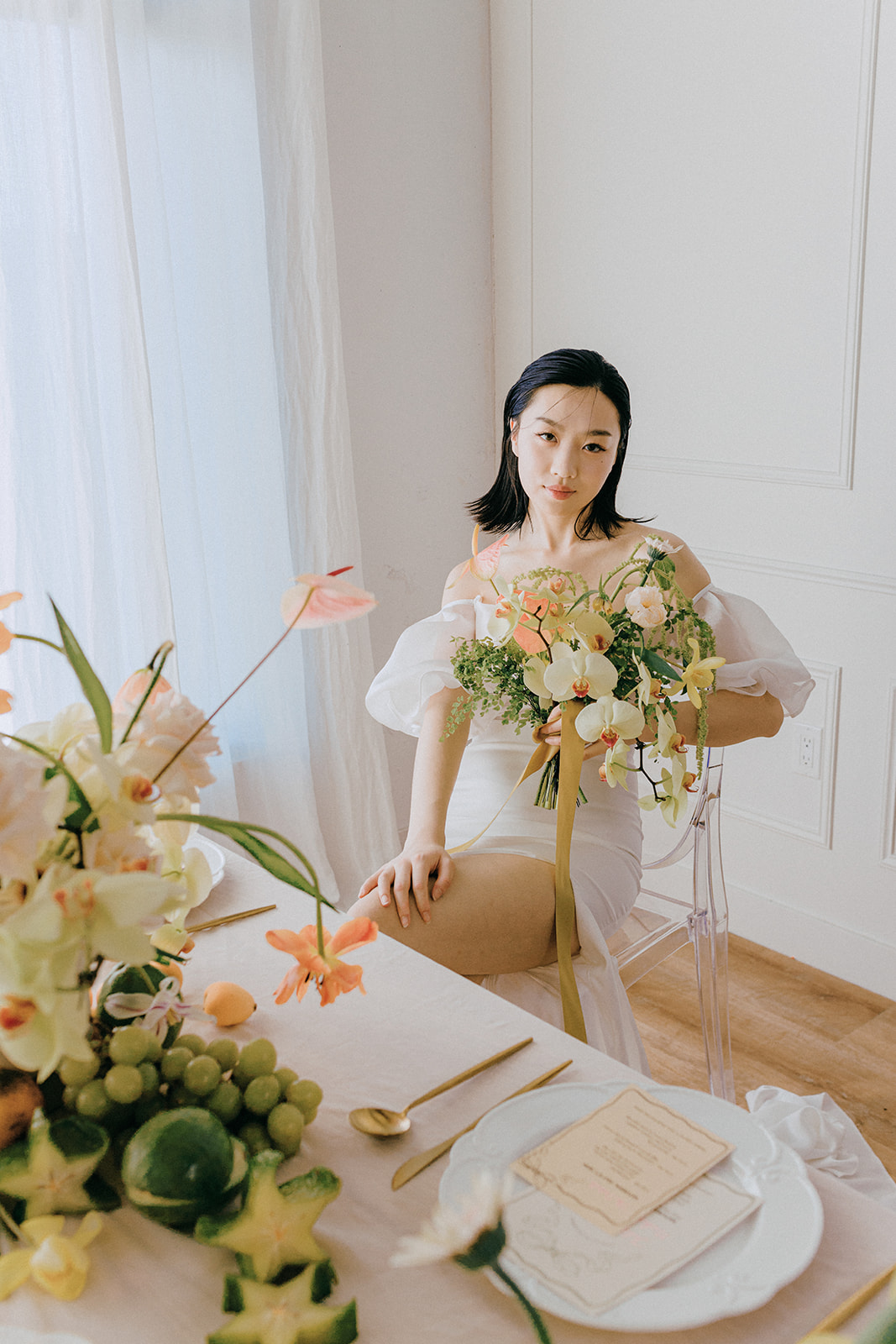 A bride in an elegant, off-the-shoulder white dress. She holds a bouquet of spring-inspired flowers, including peach-toned blooms and greenery, tied with a yellow silk ribbon. Fruit and Florals Wedding Inspiration