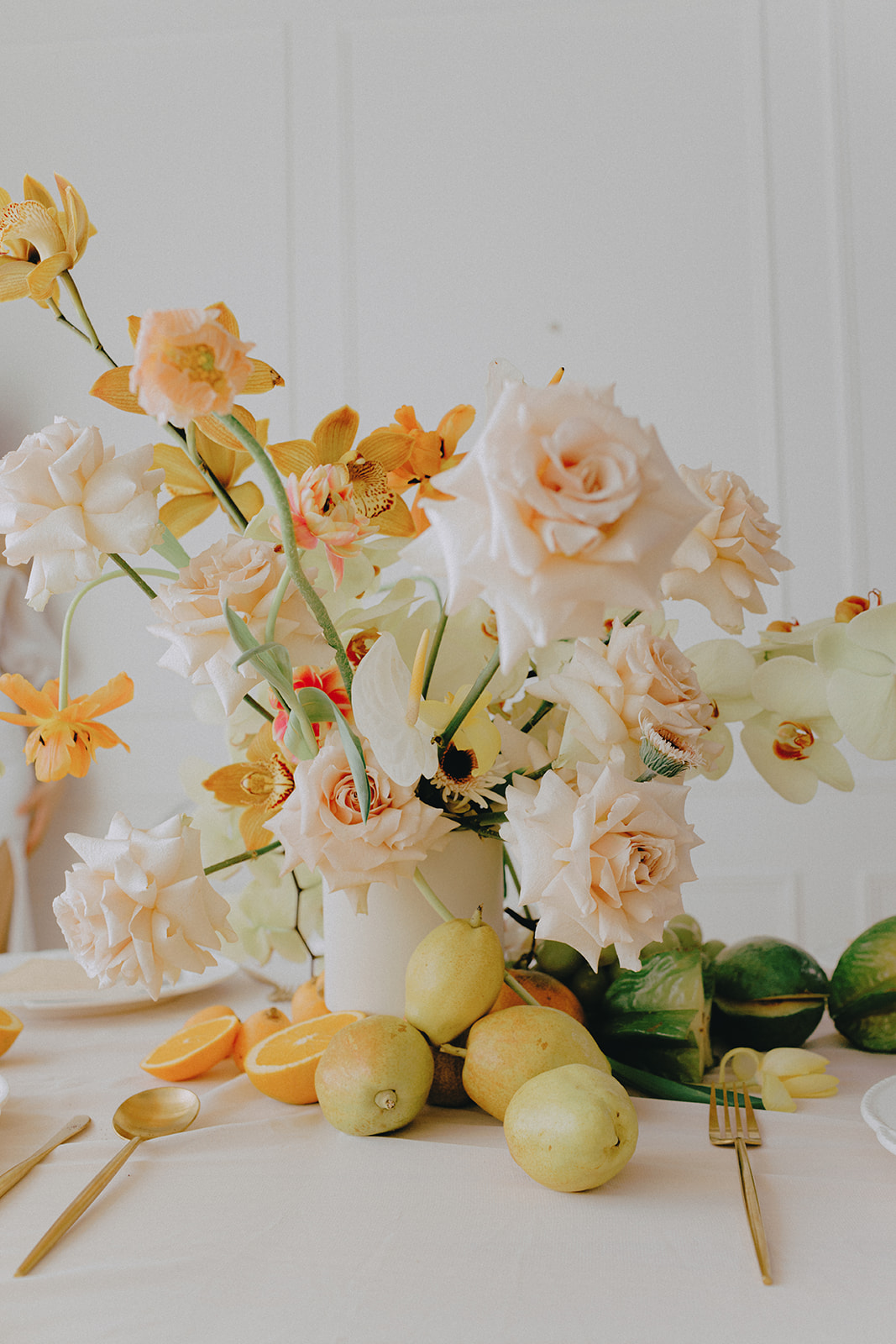 A vibrant close-up of a table arrangement featuring a lush display of flowers in soft peach, orange and cream tones. The floral arrangement is accompanied by fresh green grapes and apricots, adding a natural, elegant feel to the table setting. Citrus Fruit and Florals Wedding Centerpiece on Tablescape.