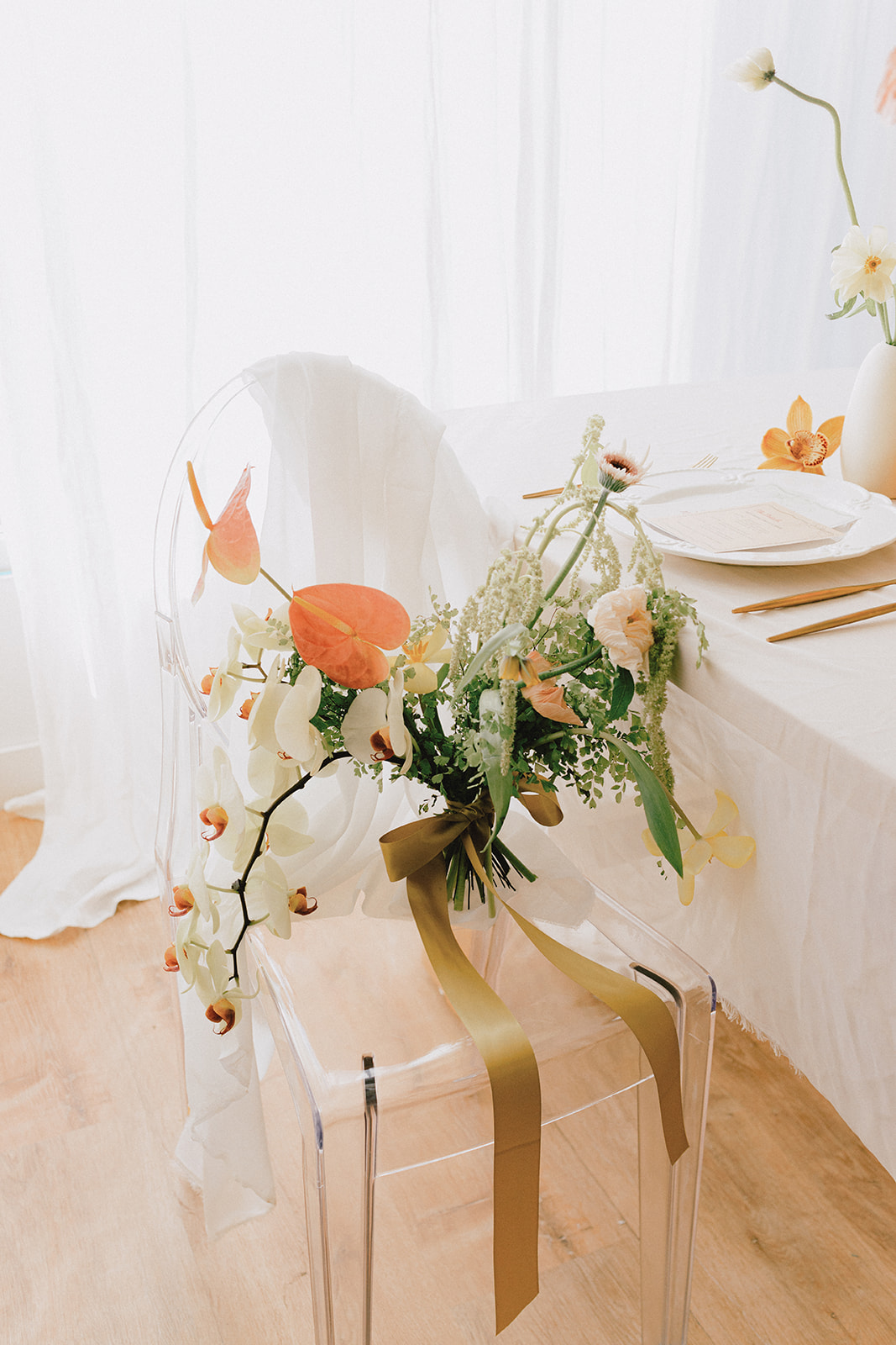 A bouquet of spring-inspired flowers, including peach-toned blooms and greenery, tied with a yellow silk ribbon on a clear glass chair. 