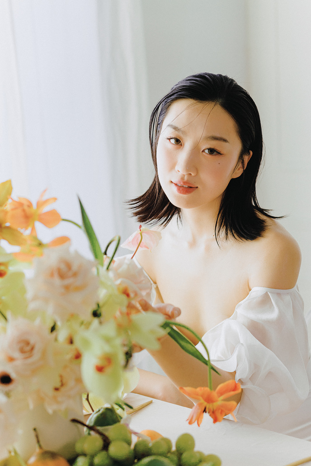 A bride in an elegant, off-the-shoulder white dress. She holds a bouquet of spring-inspired flowers, including peach-toned blooms and greenery, tied with a yellow silk ribbon.