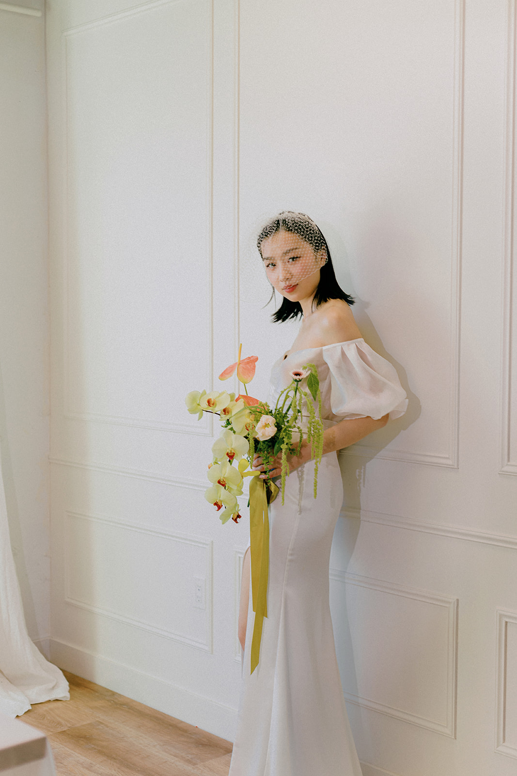 A bride in an elegant, off-the-shoulder white dress. She holds a bouquet of spring-inspired flowers, including peach-toned blooms and greenery, tied with a yellow silk ribbon.