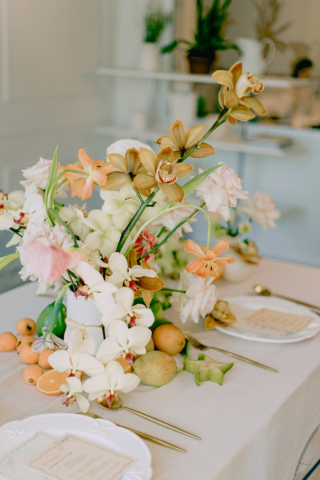 A vibrant close-up of a table arrangement featuring a lush display of flowers in soft peach, orange and cream tones. The floral arrangement is accompanied by fresh green grapes and apricots, adding a natural, elegant feel to the table setting. Fruit and Florals Wedding Table Centerpiece.