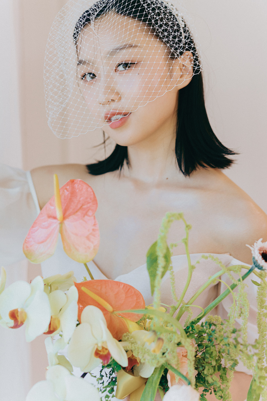 A bride in an elegant, off-the-shoulder white dress. She holds a bouquet of spring-inspired flowers, including peach-toned blooms and greenery, tied with a yellow silk ribbon.