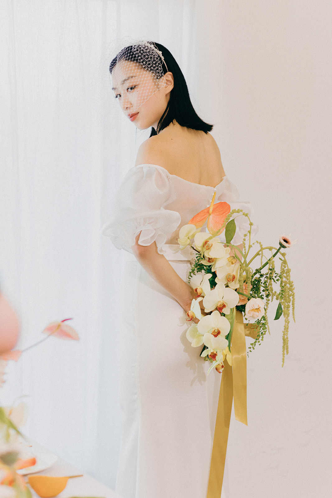 A bride in an elegant, off-the-shoulder white dress. She holds a bouquet of spring-inspired flowers, including peach-toned blooms and greenery, tied with a yellow silk ribbon. Citrus coloured wedding bouquet featured in this fresh fruit and florals wedding inspiration.