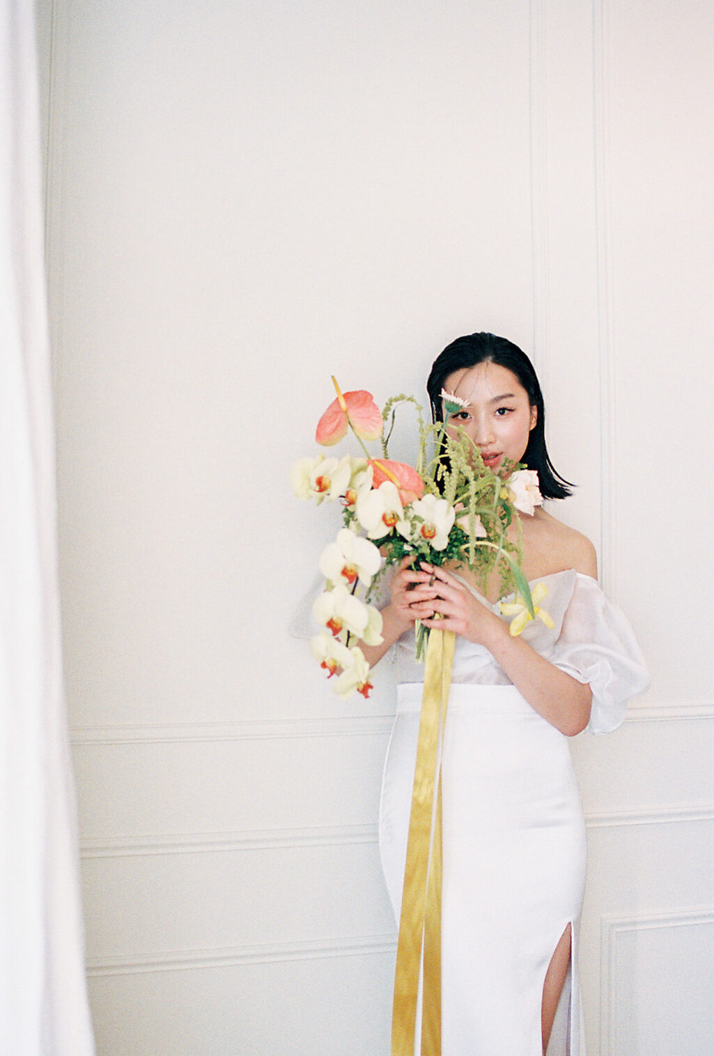 A bride in an elegant, off-the-shoulder white dress. She holds a bouquet of spring-inspired flowers, including peach-toned blooms and greenery, tied with a yellow silk ribbon. Citrus coloured wedding bouquet featured in this fresh fruit and florals wedding inspiration.