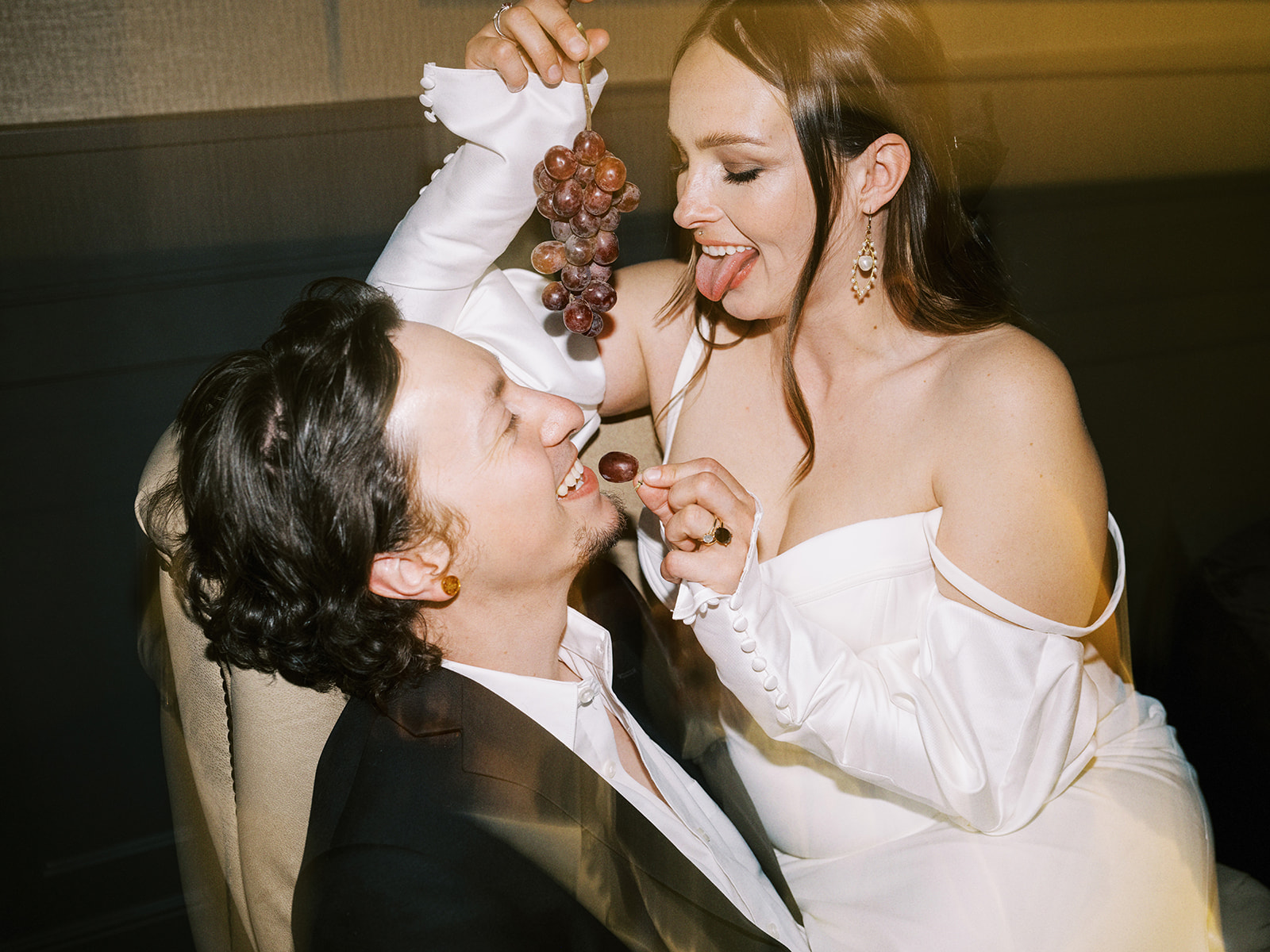 A couple in formal wedding attire shares a playful moment. The woman, in an off-shoulder white bridal gown, is teasingly holding a bunch of grapes above the groom's face, who is in a suit, while she holds a single grape towards his mouth. She smiles playfully for this unconventional wedding portrait.