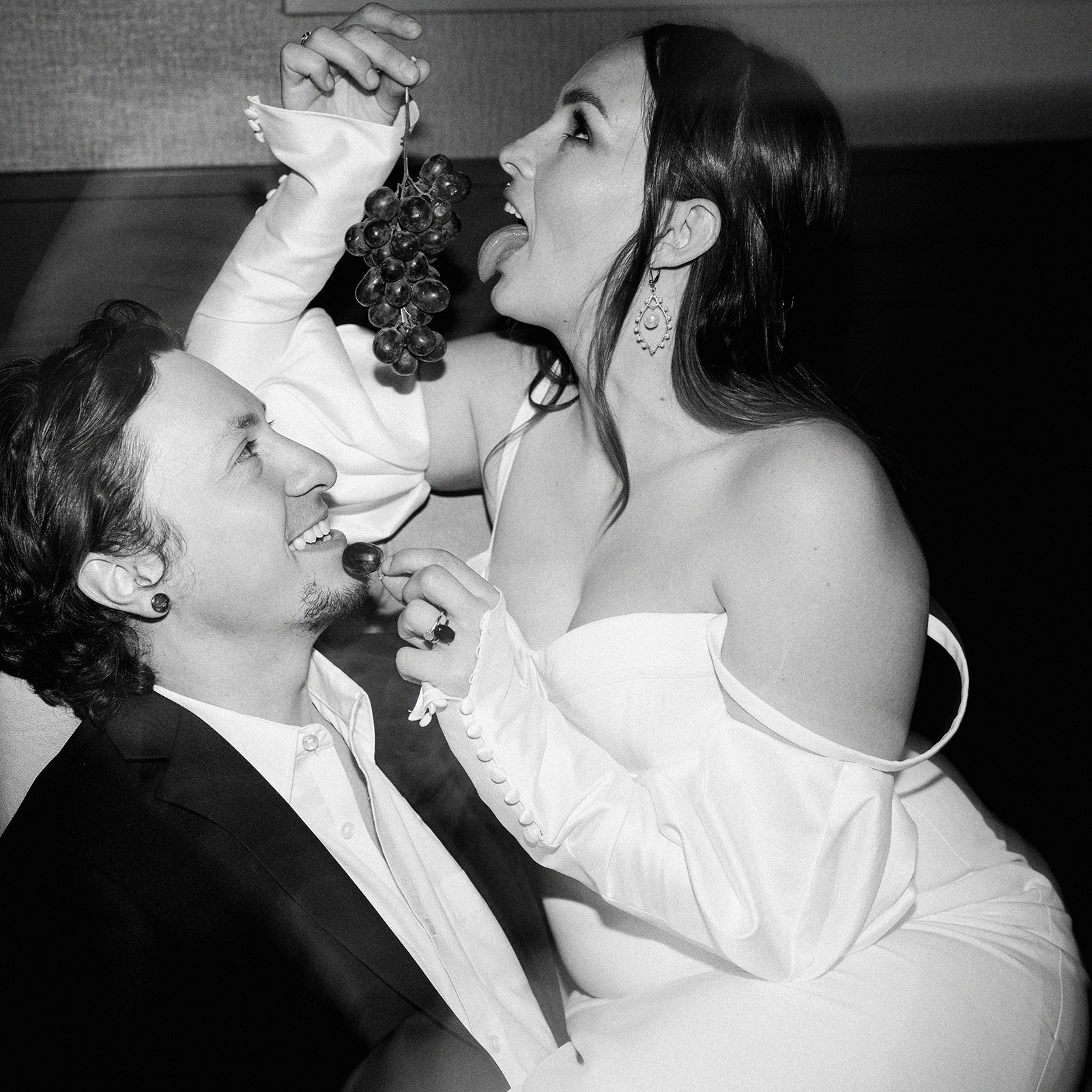 A couple in formal wedding attire shares a playful moment. The woman, in an off-shoulder white bridal gown, is teasingly holding a bunch of grapes above the groom's face, who is in a suit, while she holds a single grape towards his mouth. She smiles playfully for this unconventional wedding portrait.