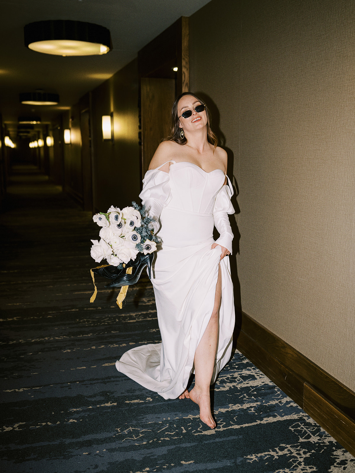 A bride in a white off-shoulder bridal gown walks barefoot down a hallway, holding a bouquet of flowers and wearing sunglasses. The hallway is dimly lit with wall lamps and has a patterned carpet. Edgy bridal inspiration for this hotel elopement.