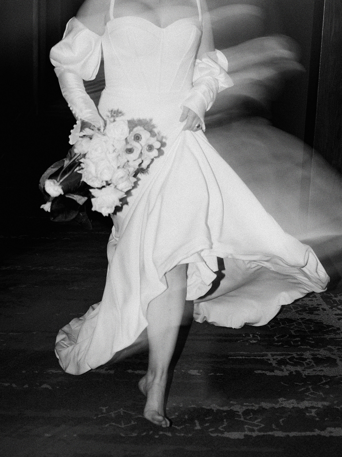 A bride in a white off-shoulder bridal gown walks barefoot down a hallway, holding a bouquet of flowers and wearing sunglasses. The hallway is dimly lit with wall lamps and has a patterned carpet. Edgy bridal inspiration for this hotel elopement.