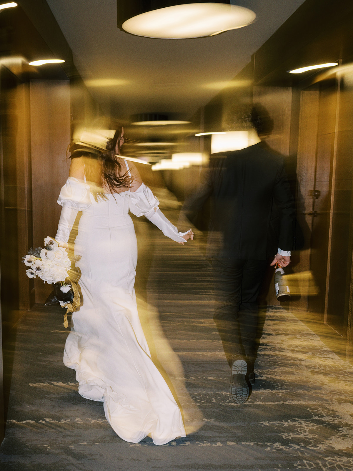 A joyful bride and groom in wedding attire holds hands, walking down a dimly lit hallway. The groom wears sunglasses and holds a bottle, while the bride holds a bouquet of white flowers, both appear relaxed and happy. Edgy wedding portrait inspiration for this hotel elopement. Unconventional Elopement Inspiration