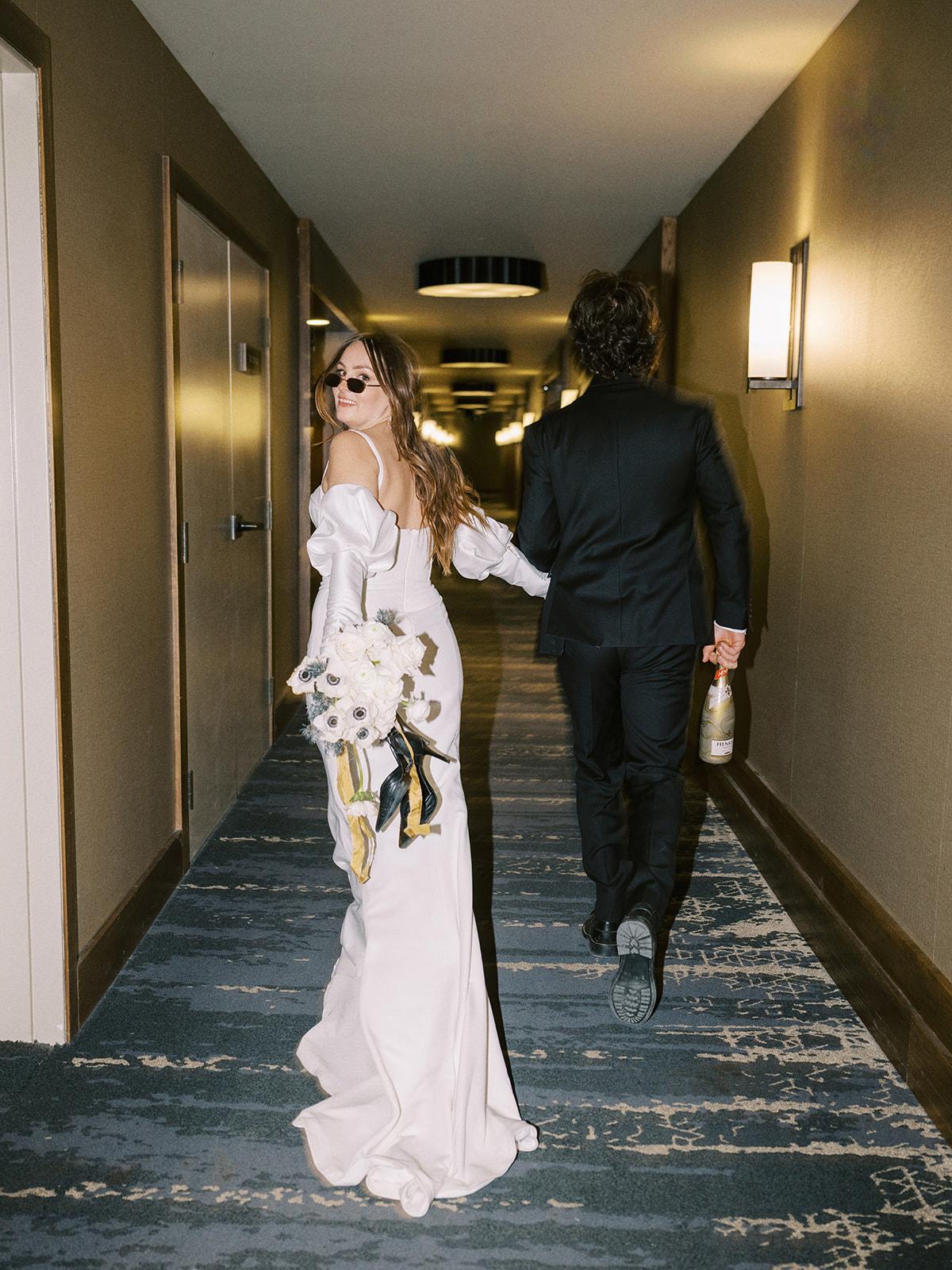 A joyful bride and groom in wedding attire holds hands, walking down a dimly lit hallway. The groom wears sunglasses and holds a bottle, while the bride holds a bouquet of white flowers, both appear relaxed and happy. Edgy wedding portrait inspiration for this hotel elopement. Unconventional Elopement Inspiration