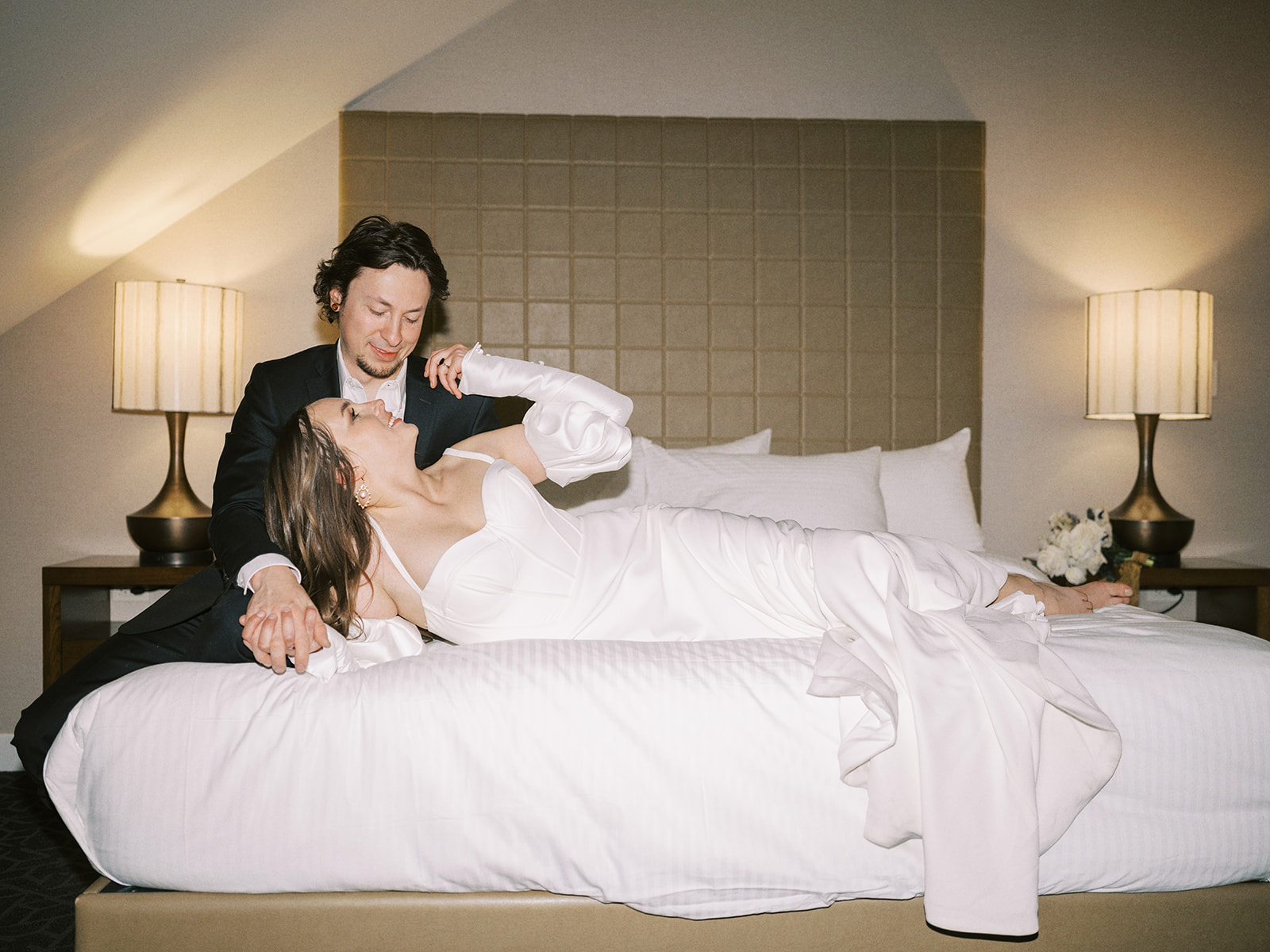 A bride and groom in formal wedding attire is embracing on a bed in a softly lit room. The woman is in a white dress, and the man is in a dark suit. They look at each other affectionately, with a lamp and padded headboard in the background. Hotel elopement wedding portraits.