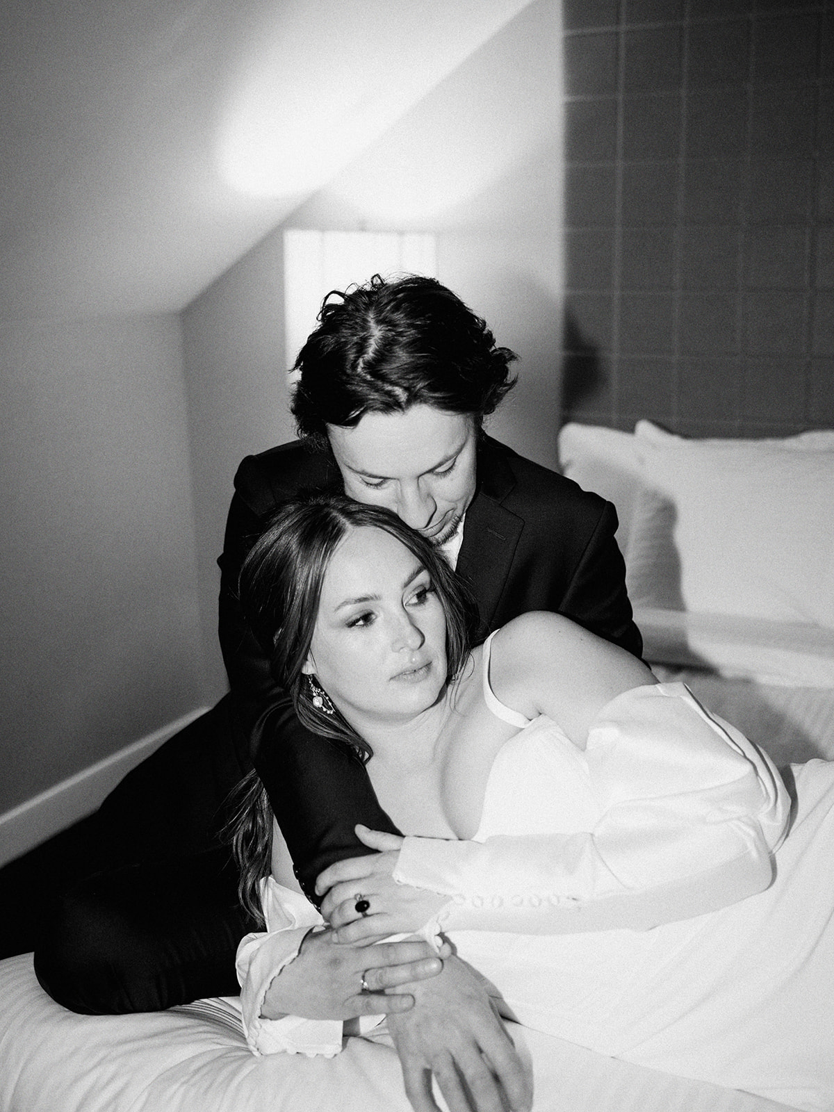 A bride and groom in formal wedding attire is embracing on a bed in a softly lit room. The woman is in a white dress, and the man is in a dark suit. They look at each other affectionately, with a lamp and padded headboard in the background. Hotel elopement wedding portraits.