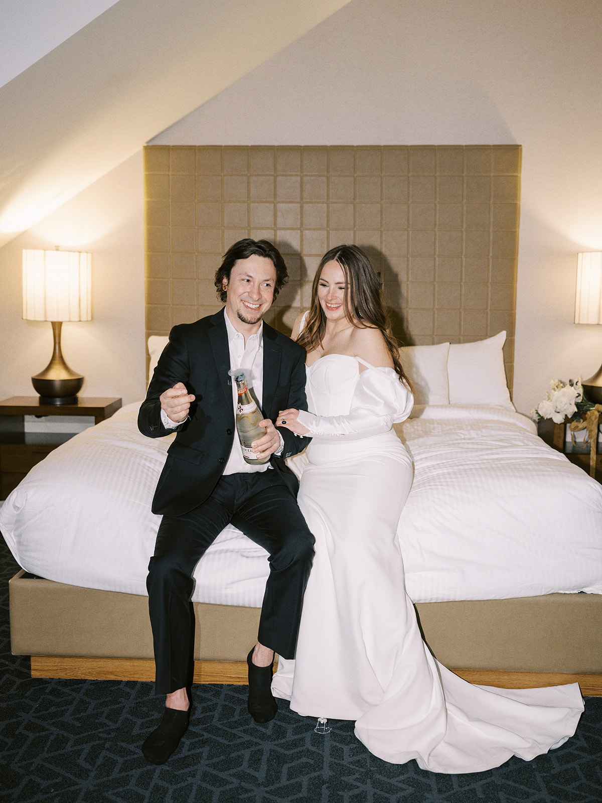 A couple in formal wedding attire sits on a hotel bed. The woman is in an off-shoulder wedding dress, holding a champagne bottle. Both are looking at each other affectionately, with soft lighting illuminating the room. Unconventional Elopement Inspiration