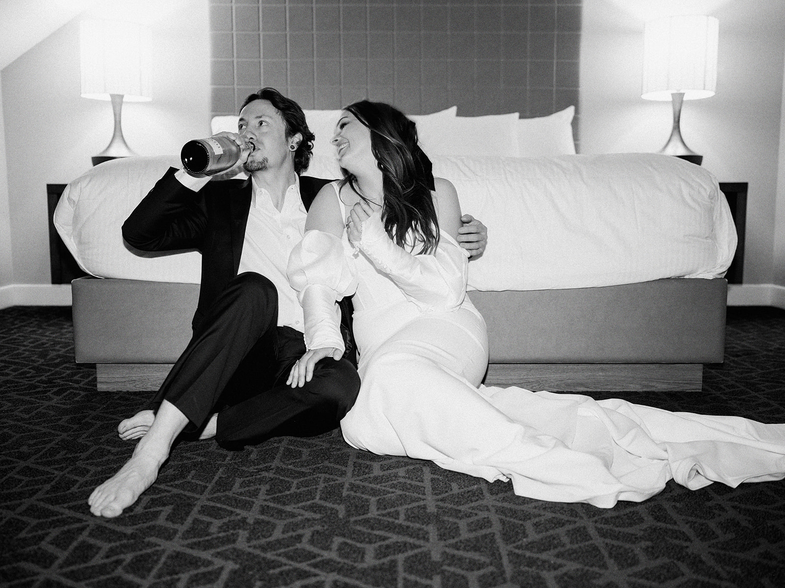 A couple, dressed in formal wedding attire, sits on the floor of a hotel room in front of a bed. The groom holds a bottle, and both appear relaxed and happy. The room is softly lit, creating a warm and intimate atmosphere. Alternative wedding edgy elopement inspiration at the Malcolm Hotel.