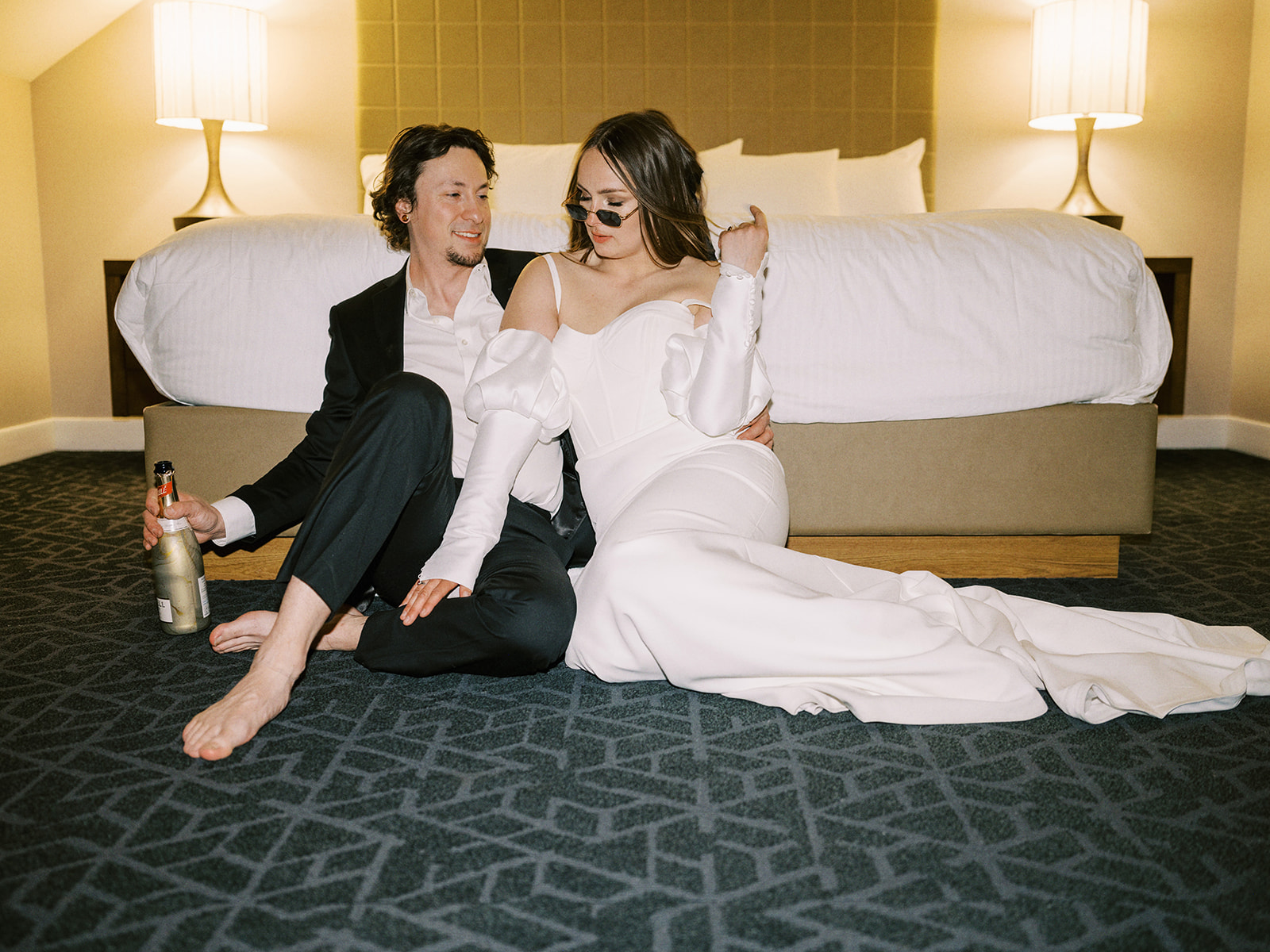 A couple, dressed in formal wedding attire, sits on the floor of a hotel room in front of a bed. The groom holds a bottle, and both appear relaxed and happy. The room is softly lit, creating a warm and intimate atmosphere. Alternative wedding edgy elopement inspiration at the Malcolm Hotel. Unconventional Elopement Inspiration