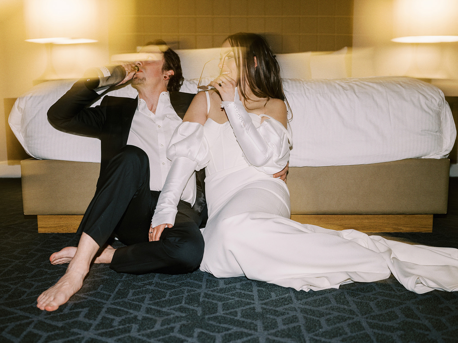 A couple, dressed in formal wedding attire, sits on the floor of a hotel room in front of a bed. The groom holds a bottle, and both appear relaxed and happy. The room is softly lit, creating a warm and intimate atmosphere. Alternative wedding edgy elopement inspiration at the Malcolm Hotel.