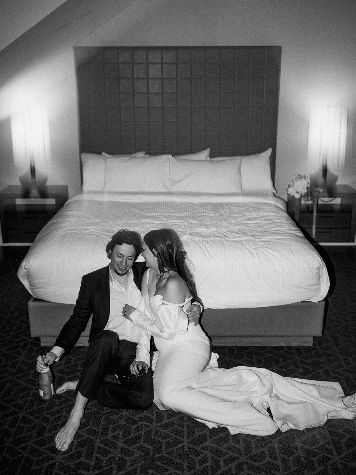 A couple, dressed in formal wedding attire, sits on the floor of a hotel room in front of a bed, embracing and sharing a kiss. The groom holds a bottle, and both appear relaxed and happy. The room is softly lit, creating a warm and intimate atmosphere. Alternative wedding edgy elopement inspiration at the Malcolm Hotel.