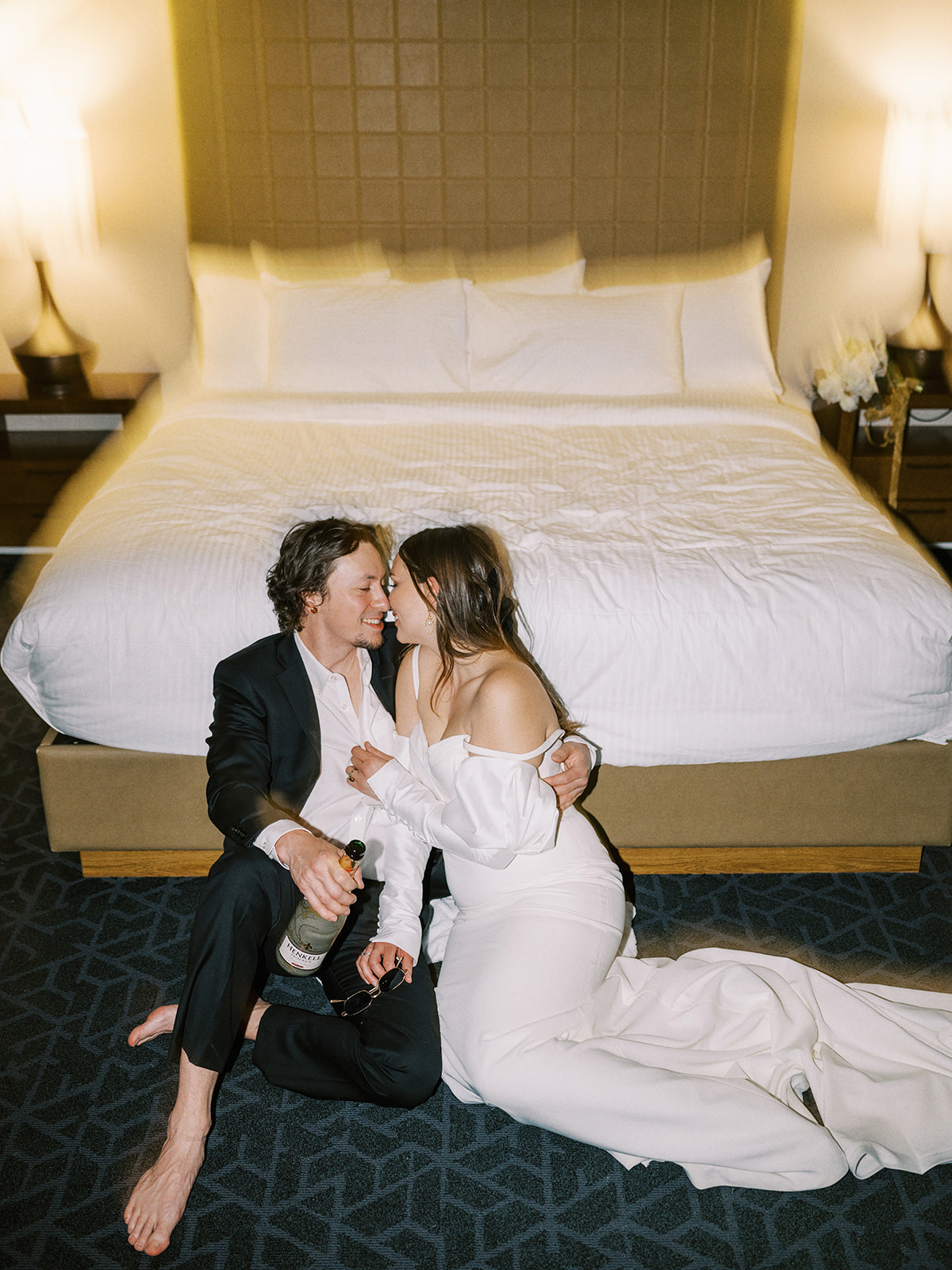 A couple, dressed in formal wedding attire, sits on the floor of a hotel room in front of a bed, embracing and sharing a kiss. The groom holds a bottle, and both appear relaxed and happy. The room is softly lit, creating a warm and intimate atmosphere. Alternative wedding edgy elopement inspiration at the Malcolm Hotel.