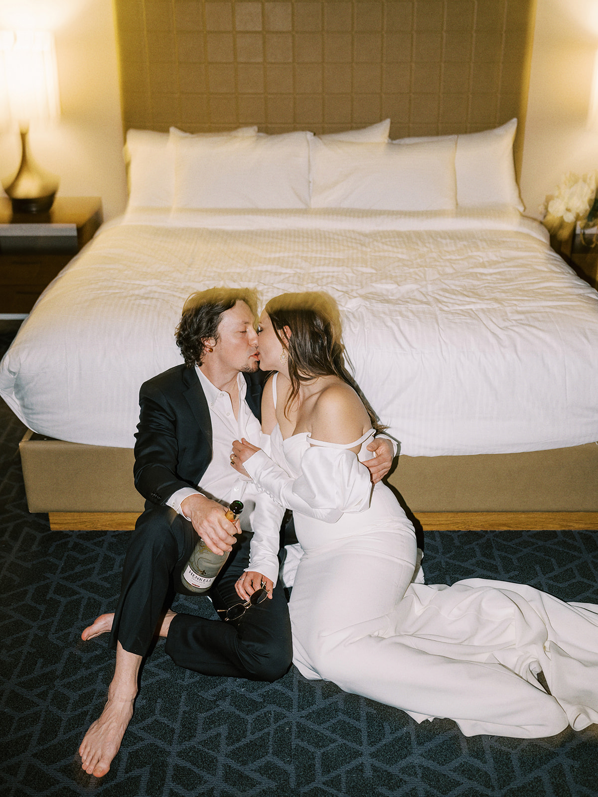 Unconventional Elopement Inspiration: A couple, dressed in formal wedding attire, sits on the floor of a hotel room in front of a bed, embracing and sharing a kiss. The groom holds a bottle, and both appear relaxed and happy. The room is softly lit, creating a warm and intimate atmosphere. Alternative wedding edgy elopement inspiration at the Malcolm Hotel.