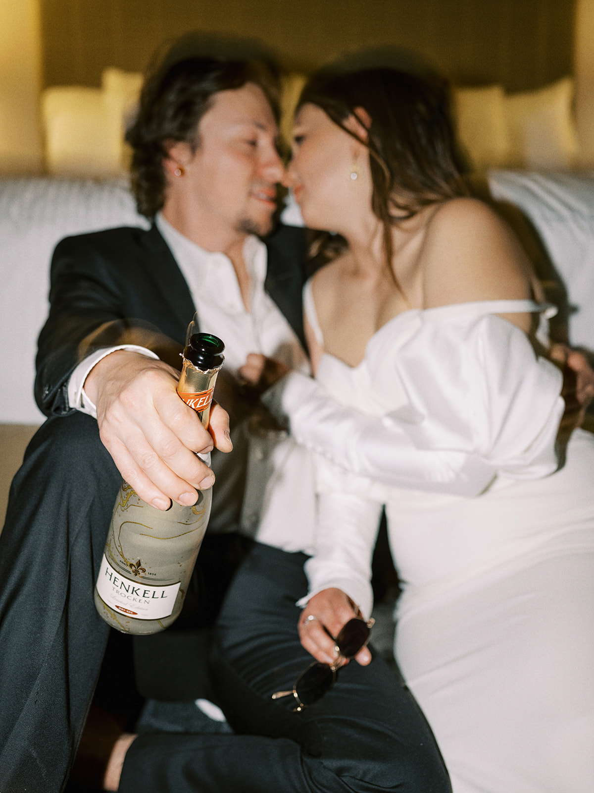 A couple, dressed in formal wedding attire, sits on the floor of a hotel room in front of a bed, embracing and sharing a kiss. The groom holds a bottle, and both appear relaxed and happy. The room is softly lit, creating a warm and intimate atmosphere. Alternative wedding edgy elopement inspiration at the Malcolm Hotel.