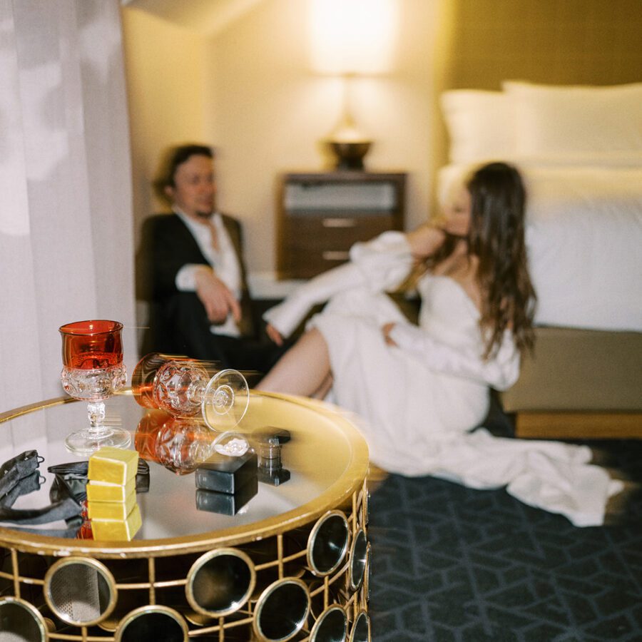 A dimly lit hotel room with a bride and groom sitting on the floor near a bed, dressed formally. In the foreground, a round table holds two glasses, a bottle, and some small boxes. The atmosphere is intimate and relaxed.