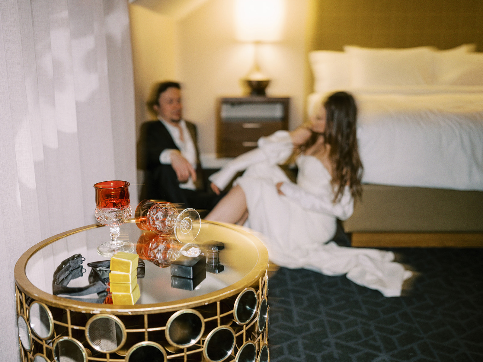 A dimly lit hotel room with a bride and groom sitting on the floor near a bed, dressed formally. In the foreground, a round table holds two glasses, a bottle, and some small boxes. The atmosphere is intimate and relaxed.