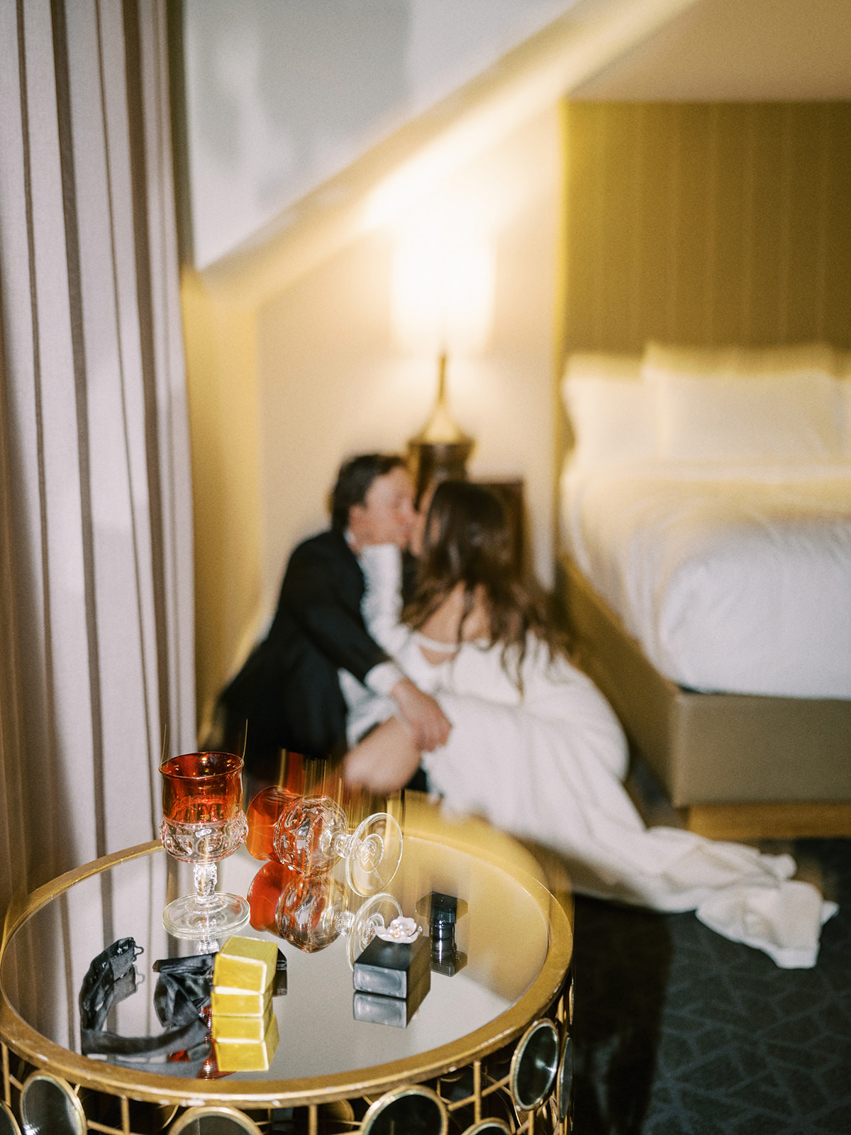 A dimly lit hotel room with a bride and groom sitting on the floor near a bed, dressed formally. In the foreground, a round table holds two glasses, a bottle, and some small boxes. The atmosphere is intimate and relaxed.