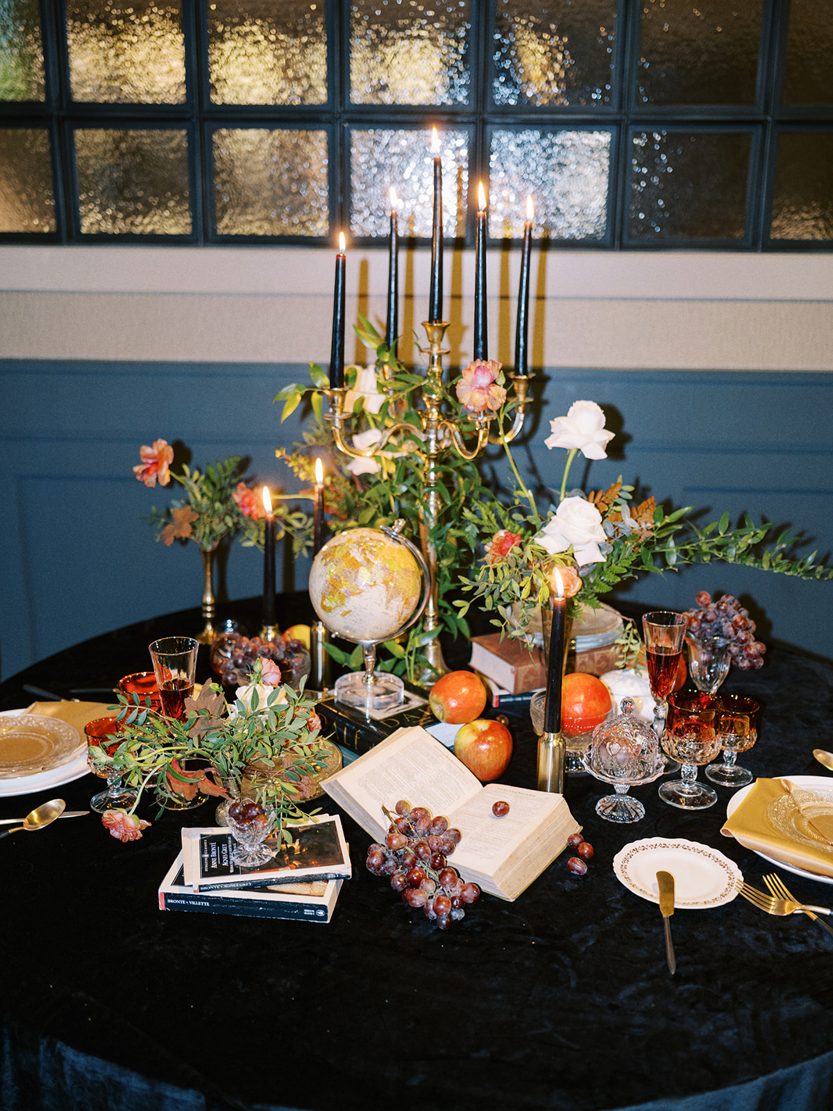 A lavishly set round table with a candelabra, open book, assorted fruits like grapes and apples, and decorative flowers. Elegant glassware and plates are arranged on a black tablecloth, creating a vintage, ornate ambiance.