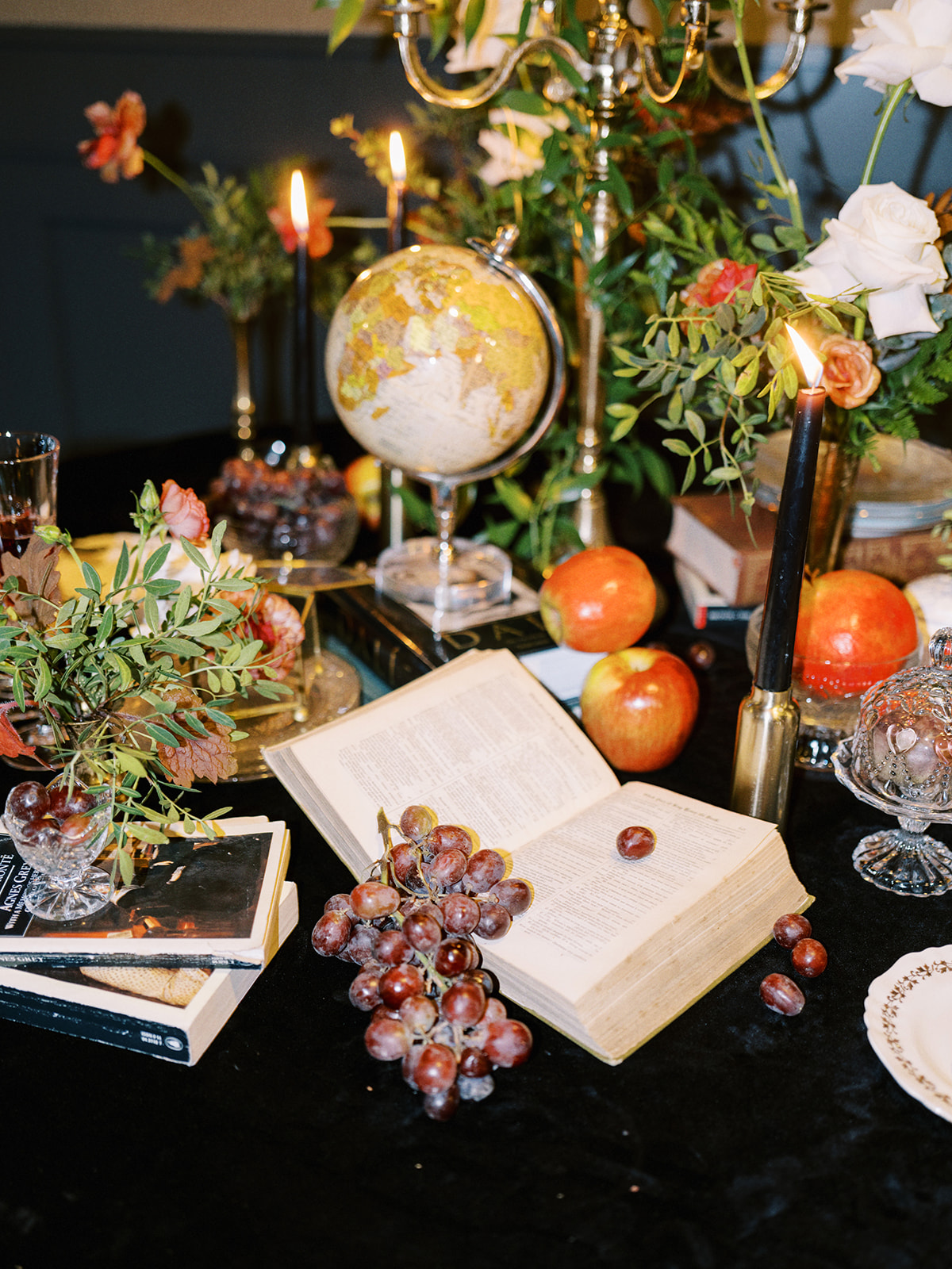 A luxurious table setting with a navy blue tablecloth, gold napkins, and crystal glassware. Black candles are lit, surrounded by floral arrangements, a small globe, and a fruit arrangement, creating an elegant, opulent atmosphere.