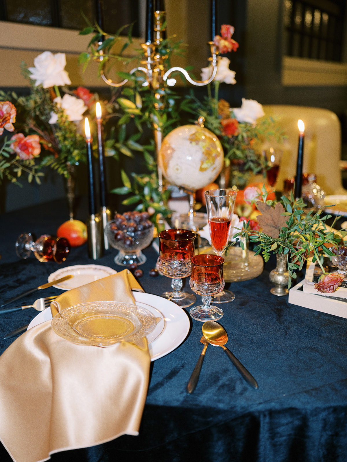 A luxurious table setting with a navy blue tablecloth, gold napkins, and crystal glassware. Black candles are lit, surrounded by floral arrangements, a small globe, and a fruit arrangement, creating an elegant, opulent atmosphere.