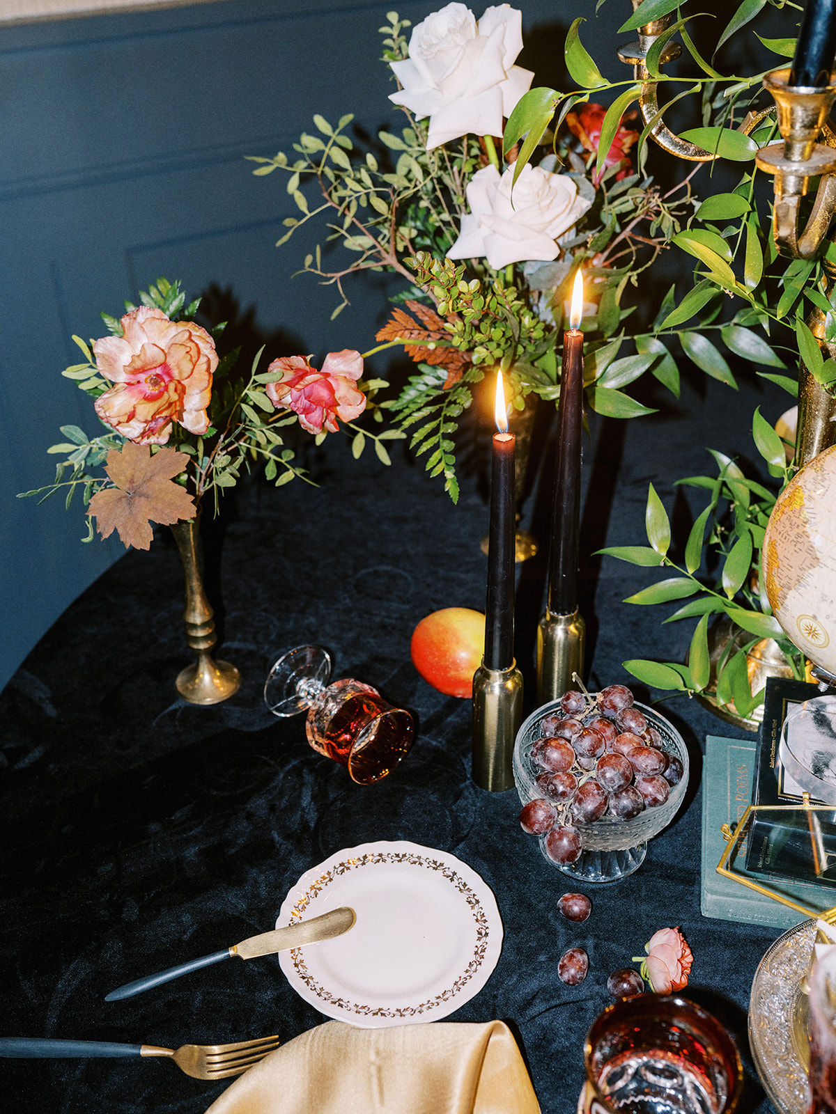 A lavishly set round table with a candelabra, open book, assorted fruits like grapes and apples, and decorative flowers. Elegant glassware and plates are arranged on a black tablecloth, creating a vintage, ornate ambiance.