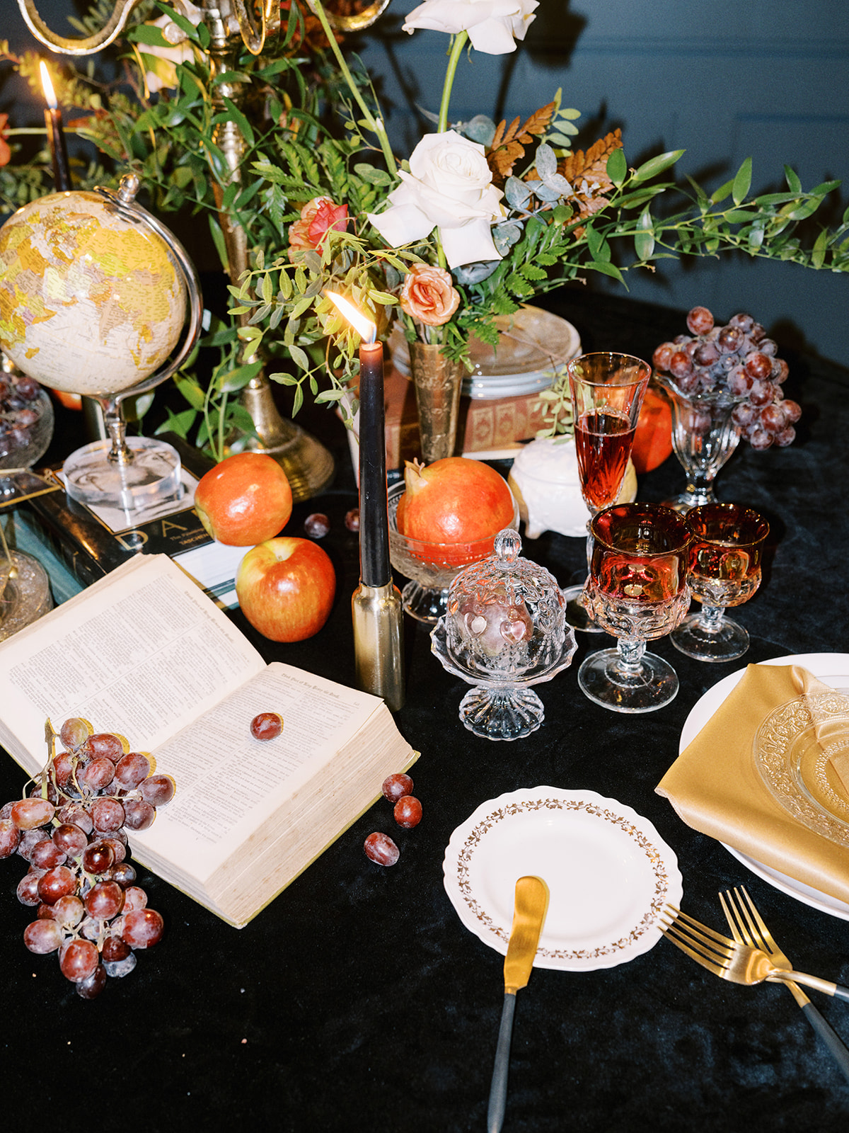 A lavishly set round table with a candelabra, open book, assorted fruits like grapes and apples, and decorative flowers. Elegant glassware and plates are arranged on a black tablecloth, creating a vintage, ornate ambiance.