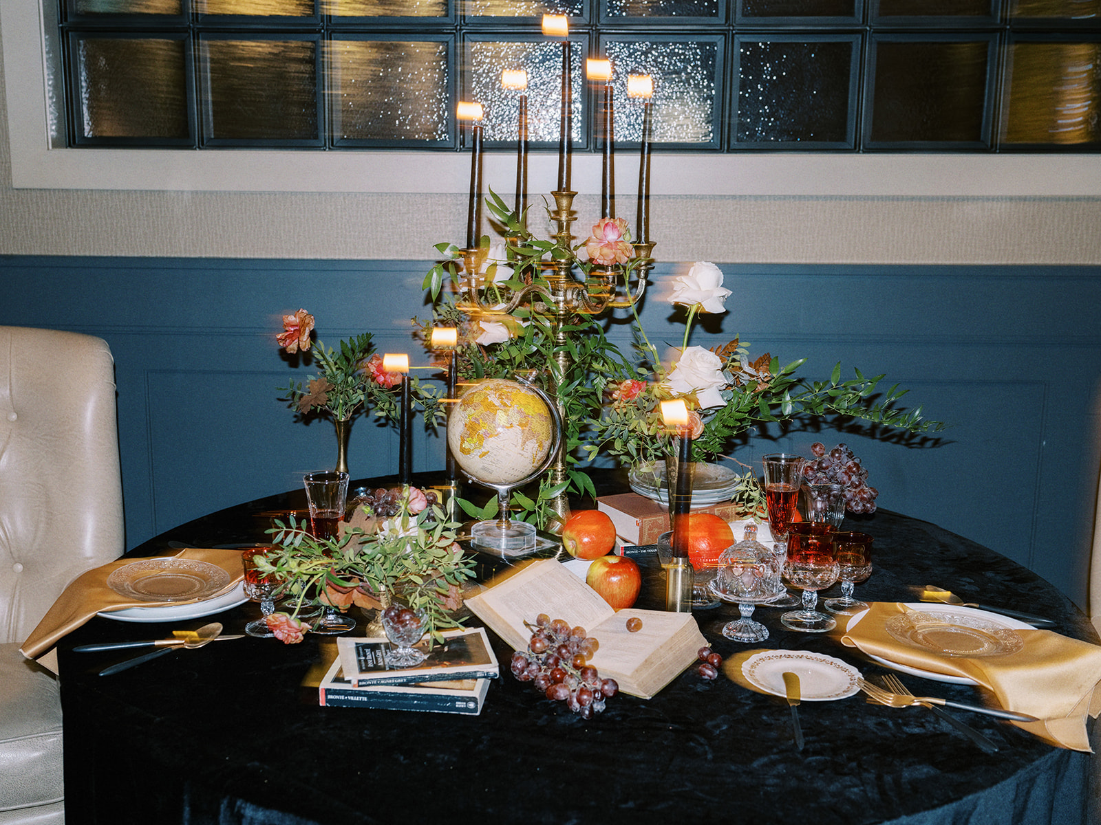 A luxurious table setting with a navy blue tablecloth, gold napkins, and crystal glassware. Black candles are lit, surrounded by floral arrangements, a small globe, and a fruit arrangement, creating an elegant, opulent atmosphere.