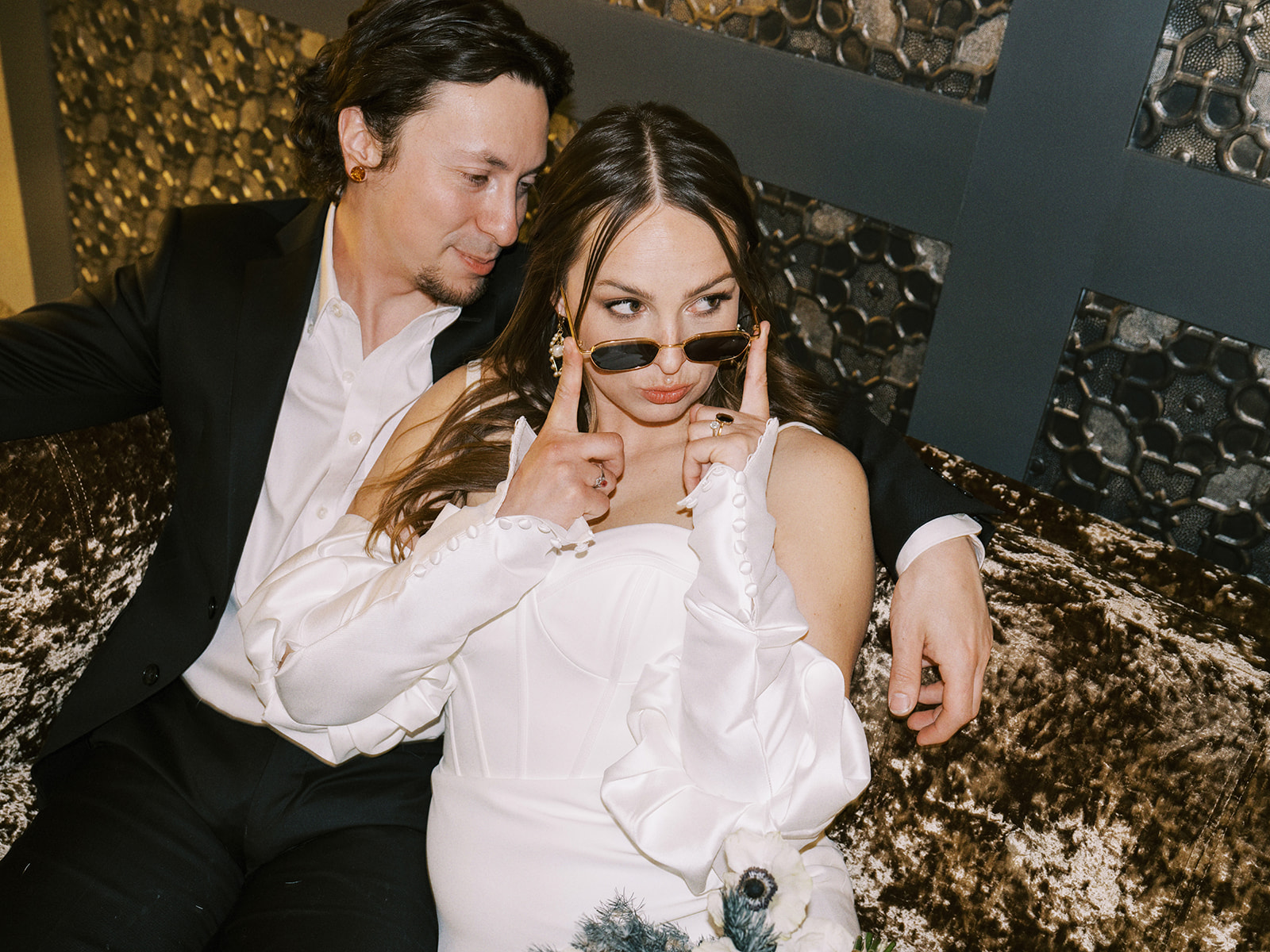 A couple sits together on a couch. The bride, wearing a white long sleeve off the shoulder bridal gown, playfully adjusts her sunglasses while smiling at the man. The man, in a black suit and white shirt, looks at her affectionately. Nontraditional wedding portraits at the Malcolm Hotel.