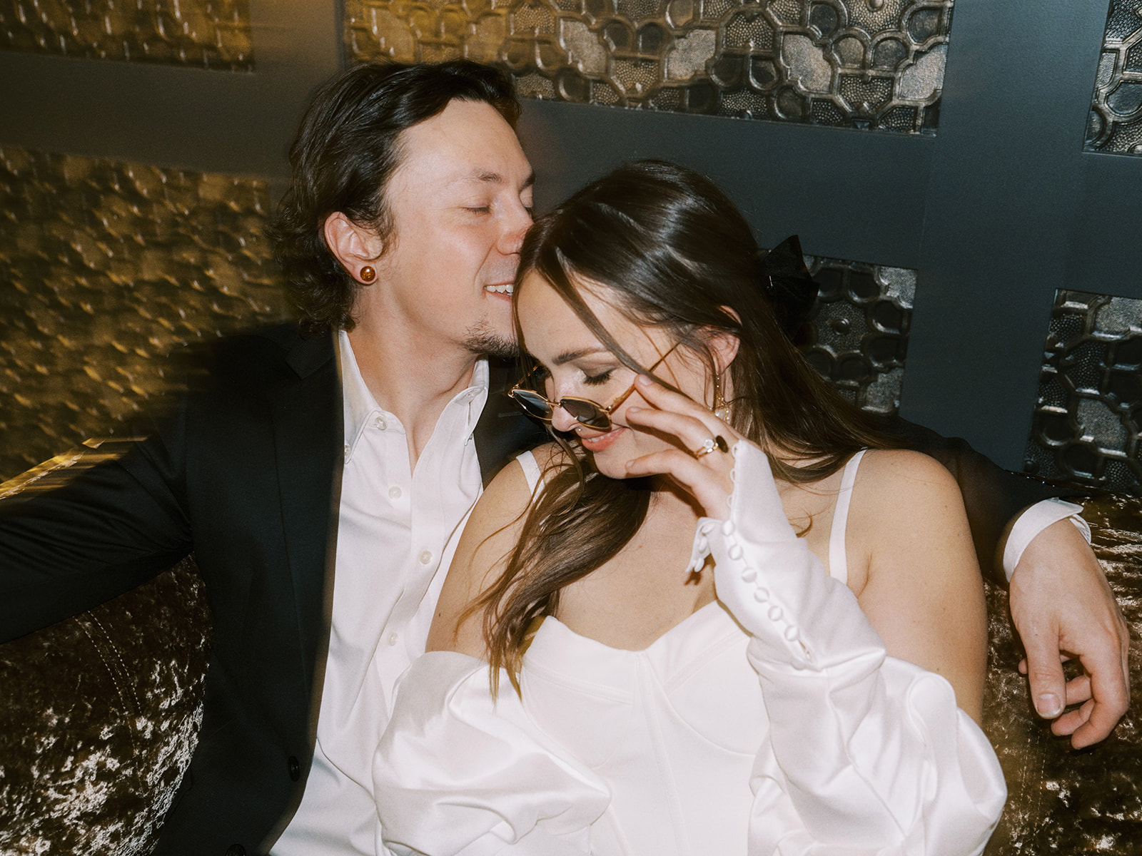 A couple sits together on a couch. The bride, wearing a white long sleeve off the shoulder bridal gown, playfully adjusts her sunglasses while smiling at the man. The man, in a black suit and white shirt, looks at her affectionately. Nontraditional wedding portraits at the Malcolm Hotel.