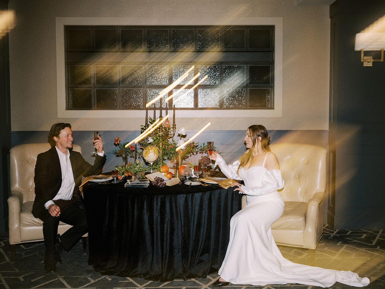 A bride and groom sit at an elegantly decorated table with a dark tablecloth, floral centerpiece, and candles. The groom wears a black suit, and the bride wears a long bridal gown with off the shoulder sleeves. They are raising glasses to each other in a dimly lit room.